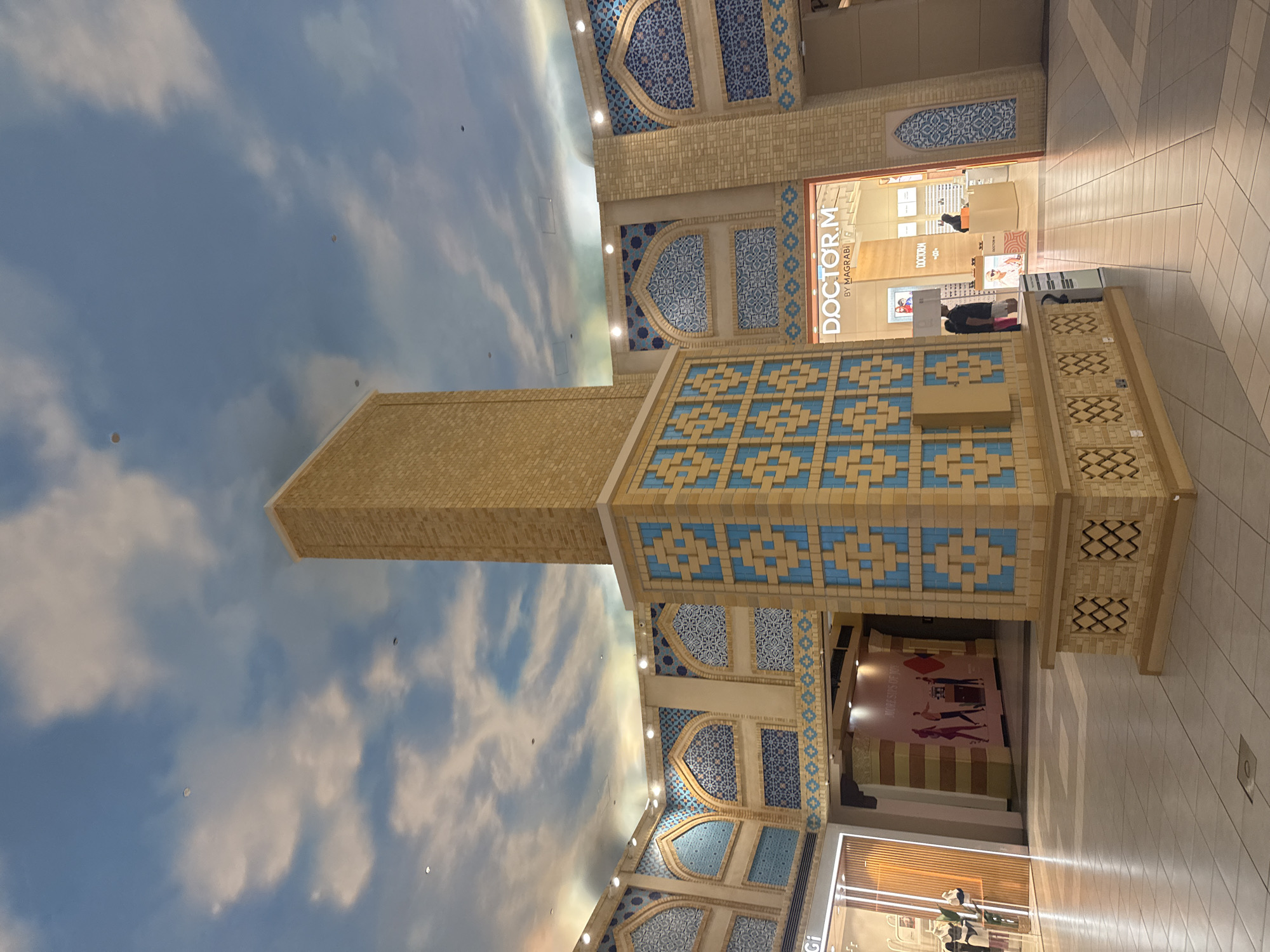 The image shows the interior of the Mall of the Emirates in Dubai, United Arab Emirates. A large, square pillar with a decorative pattern of blue and tan tiles stands in the foreground. The pillar is topped with a rectangular structure made of tan bricks, reaching towards a ceiling painted with a sky and cloud mural. The surrounding architecture features arched doorways and walls adorned with intricate blue and white geometric tile patterns. Several storefronts are visible, including "DOCTOR.M BY MAGRABI" which sells eyewear. People are seen inside the stores, one behind a counter and another browsing merchandise. The floor is tiled with light-colored square tiles. The overall impression is a well-lit, upscale indoor shopping environment with a decorative, culturally inspired design.