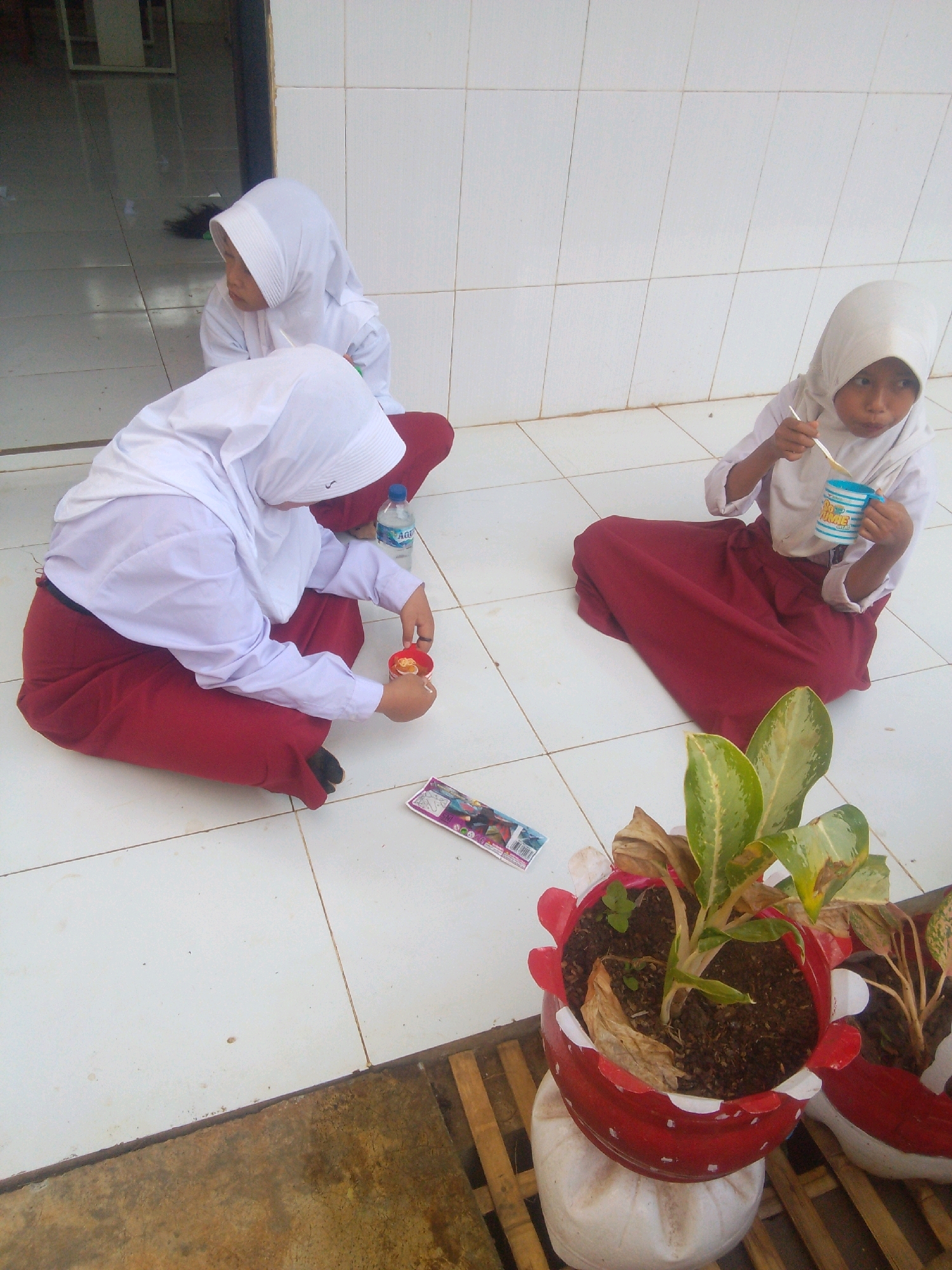 In the city of Betek, Indonesia, three young girls wearing white hijabs and maroon skirts are seated on a white tiled floor. One girl on the left is holding a small red container with a spoon, seemingly engaged in an activity with it. Another girl in the center is also seated, and a blue bottle of "Aqua" mineral water is visible beside her. The third girl on the right is eating from a blue and white striped cup with a spoon. In the foreground, a potted plant with green and yellow leaves sits in a red and white pot, resting on a wooden surface. The background consists of a tiled wall and a doorway leading to an interior space.