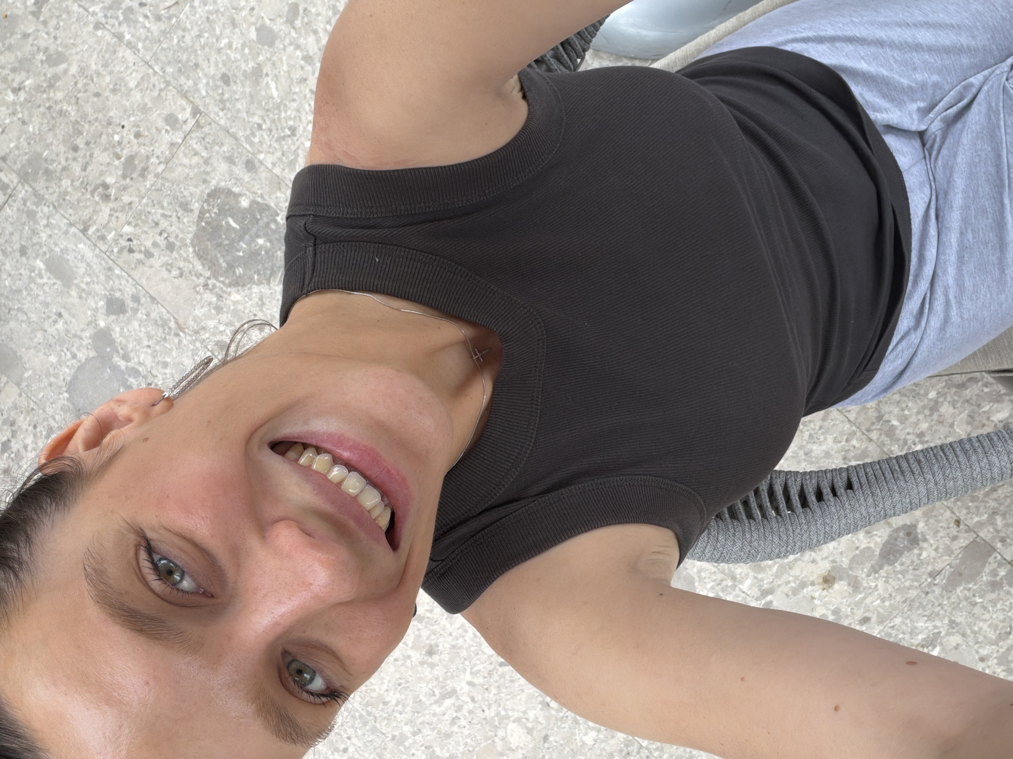 A young adult female is captured in a close-up, upward-facing selfie, smiling broadly directly at the camera. She appears to be reclining or lying down, with her head slightly tilted. Her expression conveys happiness and warmth, with her eyes open and engaging. She has light-colored eyes and a wide smile, revealing her teeth.
She is wearing a dark, likely charcoal or black, ribbed short-sleeved top. A thin silver necklace with a small cross pendant is visible around her neck, and a silver earring can be seen in her left ear. Her hair is mostly pulled back. Her lower body is clad in light grey casual trousers, possibly sweatpants.
The background consists of a light grey, speckled tiled floor, suggesting an outdoor patio or balcony. Partially visible behind her is a grey, woven outdoor chair or lounge surface on which she appears to be resting. The lighting is bright and natural, indicating it is daytime. The scene suggests a relaxed, casual moment in an outdoor setting, potentially in Budva, Montenegro. No visible text is present in the image.