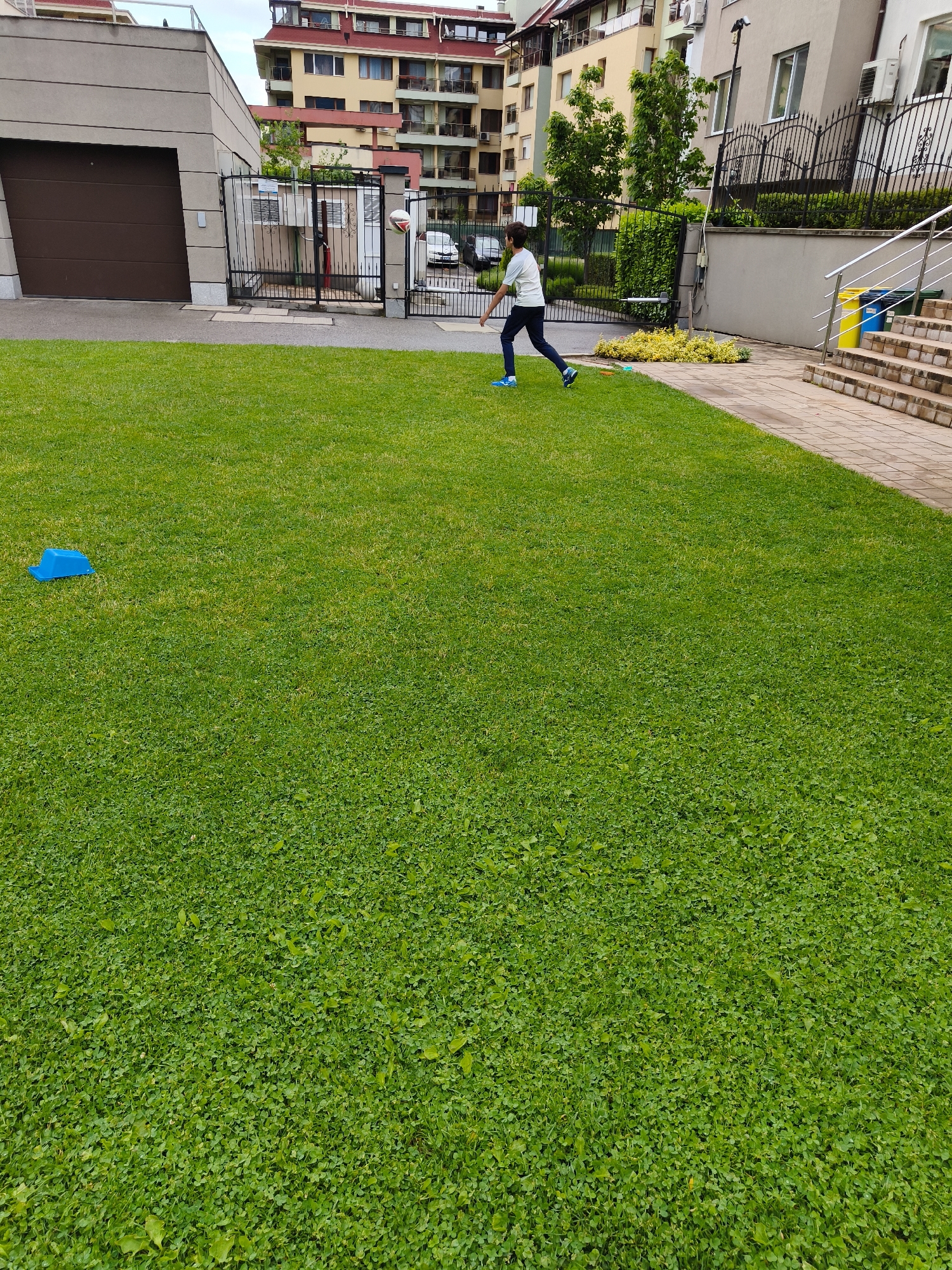 This image captures a vibrant outdoor scene in a residential area of Sofia, Bulgaria, featuring a young person engaged in play on a green lawn. The central subject is a boy, dressed in a white t-shirt, dark pants, and blue sneakers, running actively across the midground of the lawn from left to right. He appears to be playing a game, with a small, white and red ball visible in the air slightly to the right of the left gate.
The foreground and midground are dominated by a lush green lawn, primarily composed of grass and clover. In the left foreground, a bright blue plastic object, possibly a game marker, is placed on the grass. Behind the lawn, a paved path borders several residential structures. On the left, a modern building with a dark brown, slanted garage door is visible, next to a black metal gate. Further back, multi-story apartment buildings with balconies and varied architectural styles, including red-tiled roofs, form the backdrop.
On the right side of the image, a set of outdoor stairs with silver metal railings leads up to a building entrance, where yellow and blue waste bins are visible. The sky is overcast, indicating a daytime setting with soft, even lighting and no harsh shadows. The overall impression is a casual, active moment in a well-maintained urban green space. No legible text is visible within the frame.