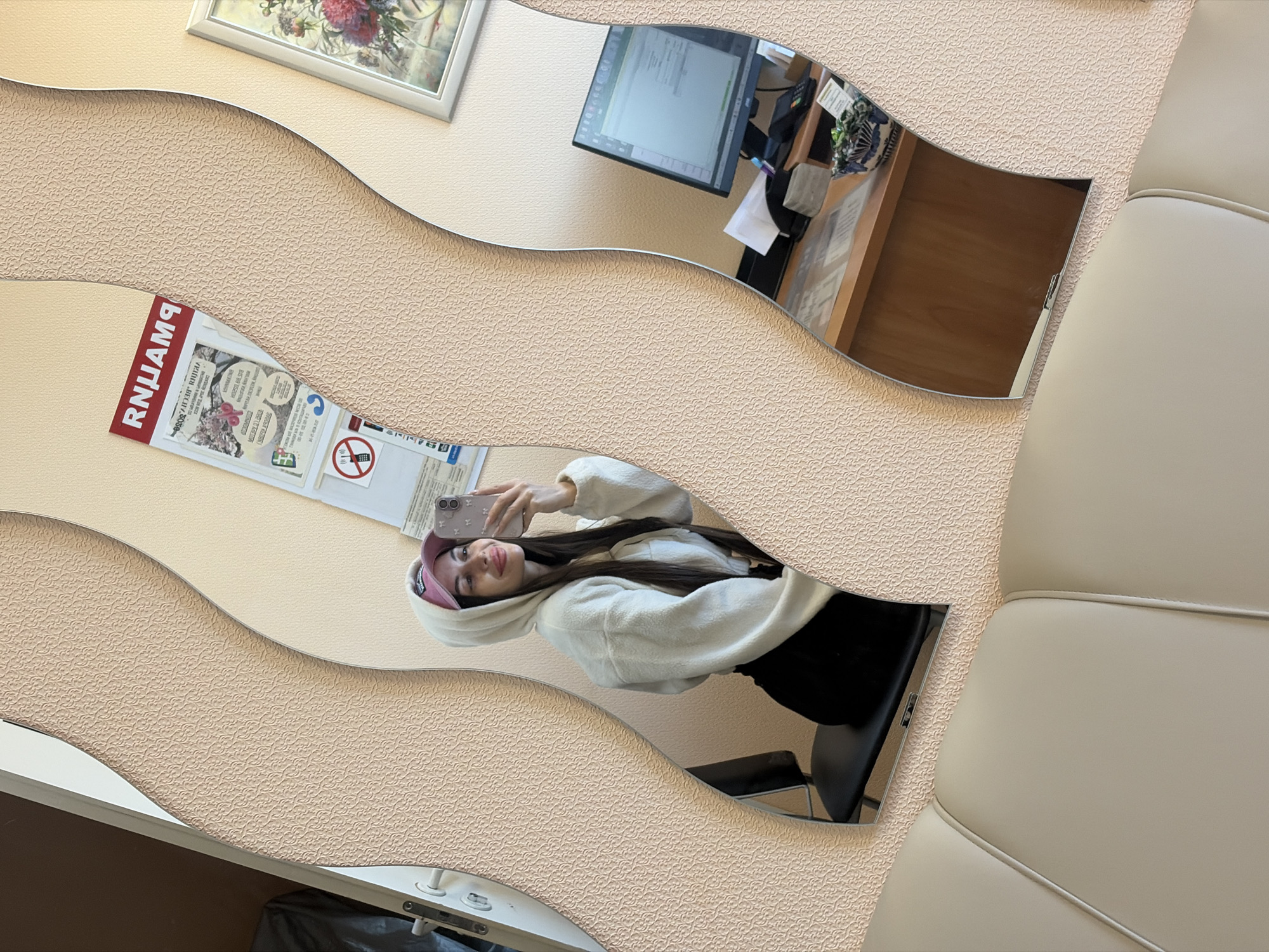 An indoor scene is depicted through a series of decorative, wavy mirrors, showing reflections of the surrounding environment and a person. The image is taken at a 90-degree rotation.
In the center, reflected in one of the wavy mirror segments, a young woman is visible, taking a selfie with a light-colored smartphone. She has long dark hair, is wearing a white fluffy hoodie, black bottoms, and a pink baseball cap. She is smiling faintly.
The background wall has a light beige, textured wallpaper with a repeating pattern. To the right, a light-colored, upholstered seating area, possibly a sofa or bench, is partially visible.
Other reflections in the mirrors include:
* A computer monitor displaying what appears to be a spreadsheet or document, positioned on a wooden desk.
* A black office chair.
* A small framed picture with floral art, hanging on the wall.
* A poster or sign, also reflected and reversed, featuring Cyrillic text. The prominent red text at the top, when reversed, clearly reads "РЕКЛАМА" (REKLAMA), meaning "ADVERTISEMENT." Below this, other smaller Cyrillic texts are visible, including phrases like "ПОРЯДОК ВЫДАЧИ КАРТЫ СКИДОК" (PROCEDURE FOR ISSUING DISCOUNT CARDS) and "СПАСИБО" (THANK YOU). A "no phone" symbol (red circle with a phone and a diagonal line) is also on the poster.
* A dark object or trash bag is partially reflected in the bottom left mirror segment.
The setting appears to be a public indoor space, such as a waiting room, office, or reception area, likely in Saint Petersburg, Russia, given the Cyrillic text. The overall atmosphere is neutral, suggesting a typical daytime indoor environment.