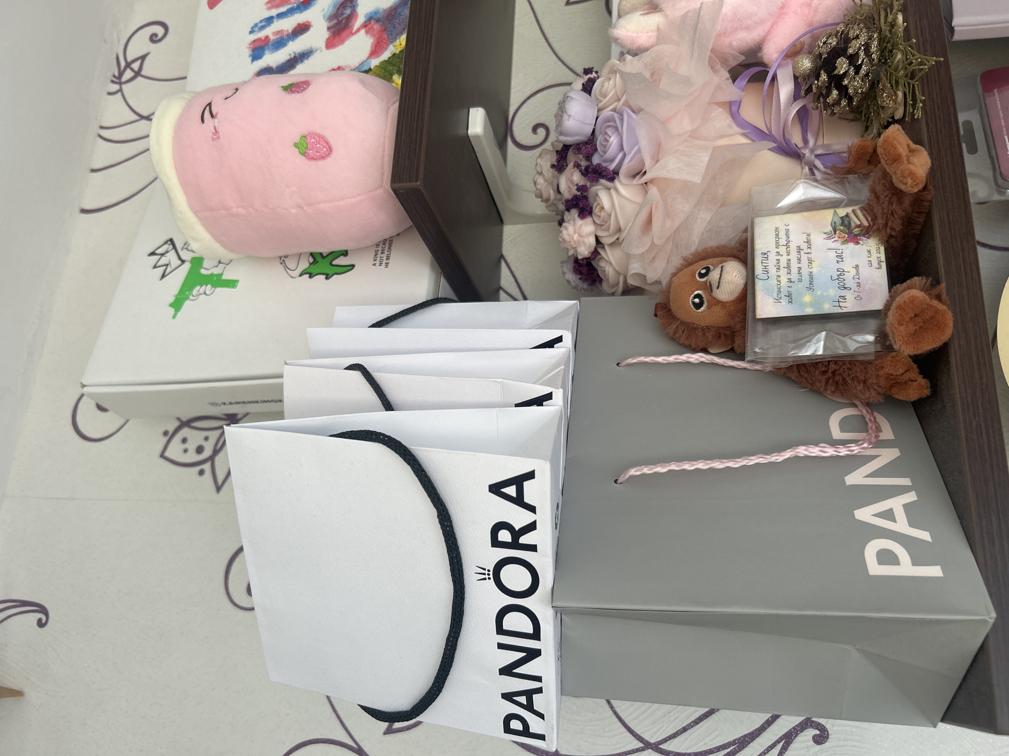 The image presents a close-up view of an arrangement on what appears to be a patterned surface, likely a table or shelf. The central focus is on several shopping bags, prominently displaying the brand "PANDORA" in bold, black lettering on a white background and a gray bag. The bags are arranged in a way that obscures some of their contents, but their presence suggests recent shopping or gifts.
Beside the bags, there is a collection of items including a plush pink toy resembling a strawberry, a bouquet of flowers in pastel shades (roses), a small teddy bear, and a gift tag. The gift tag is notable for containing text in Bulgarian, including the word "Синтия," which may be a name, and congratulatory messages like "На добър час!" (Good luck!).
The setting is domestic.