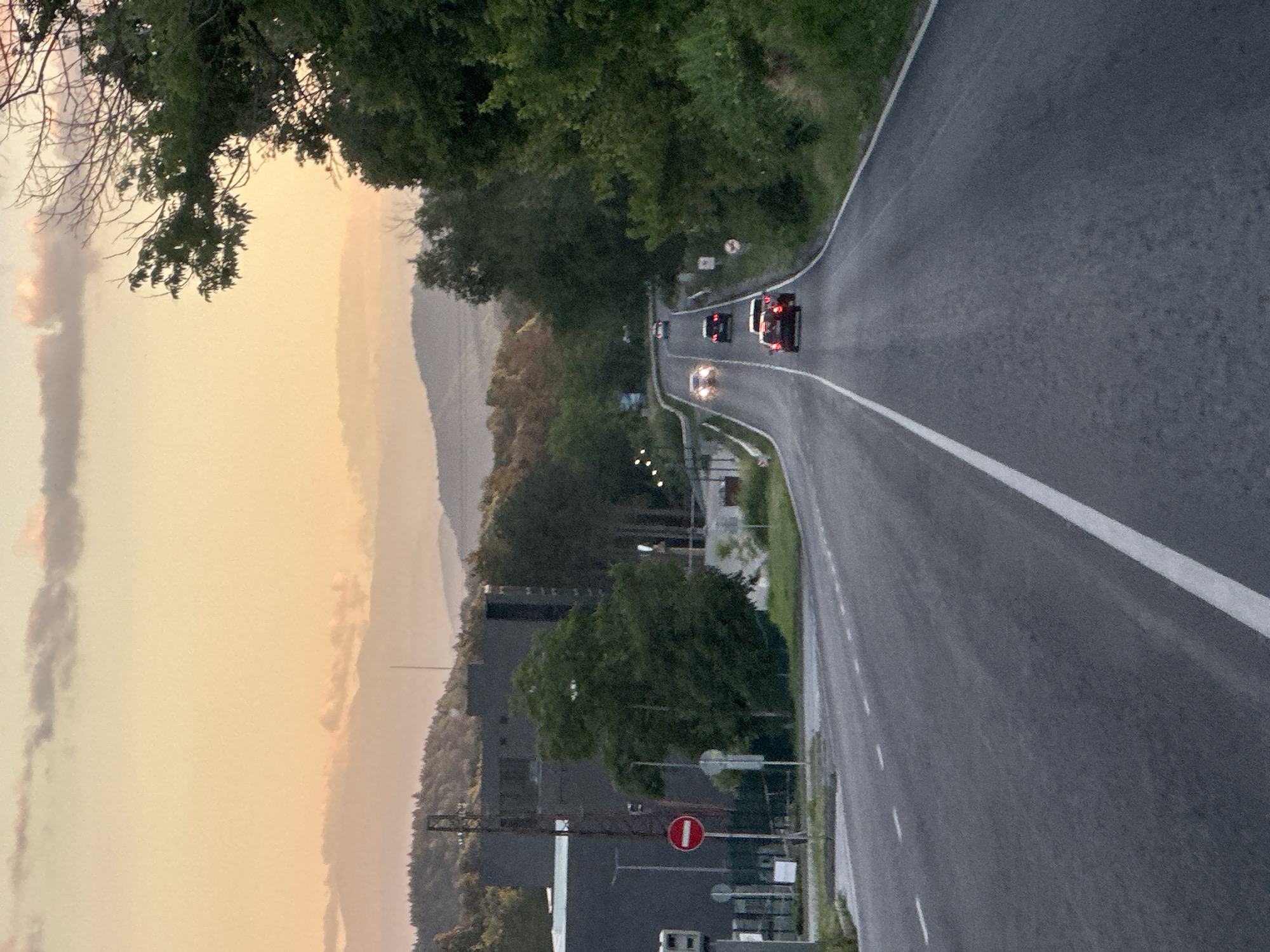 The image depicts a road with vehicles on it, likely in the vicinity of Sheremetia, Bulgaria, during what appears to be sunset. The sky is a pale orange and yellow with wispy clouds. In the foreground, the asphalt road curves to the right, marked with white lines. Several cars are visible on the road, moving away from the viewer. On the left side of the road, there's a dark building with some infrastructure, possibly an industrial facility, and a traffic sign that appears to be a "Do Not Enter" symbol. Lush green trees and vegetation line the right side of the road, with more distant hills or mountains visible in the background. The overall impression is that of a road leading out of or into a less urbanized area, with the natural landscape and the sky suggesting the end of the day.