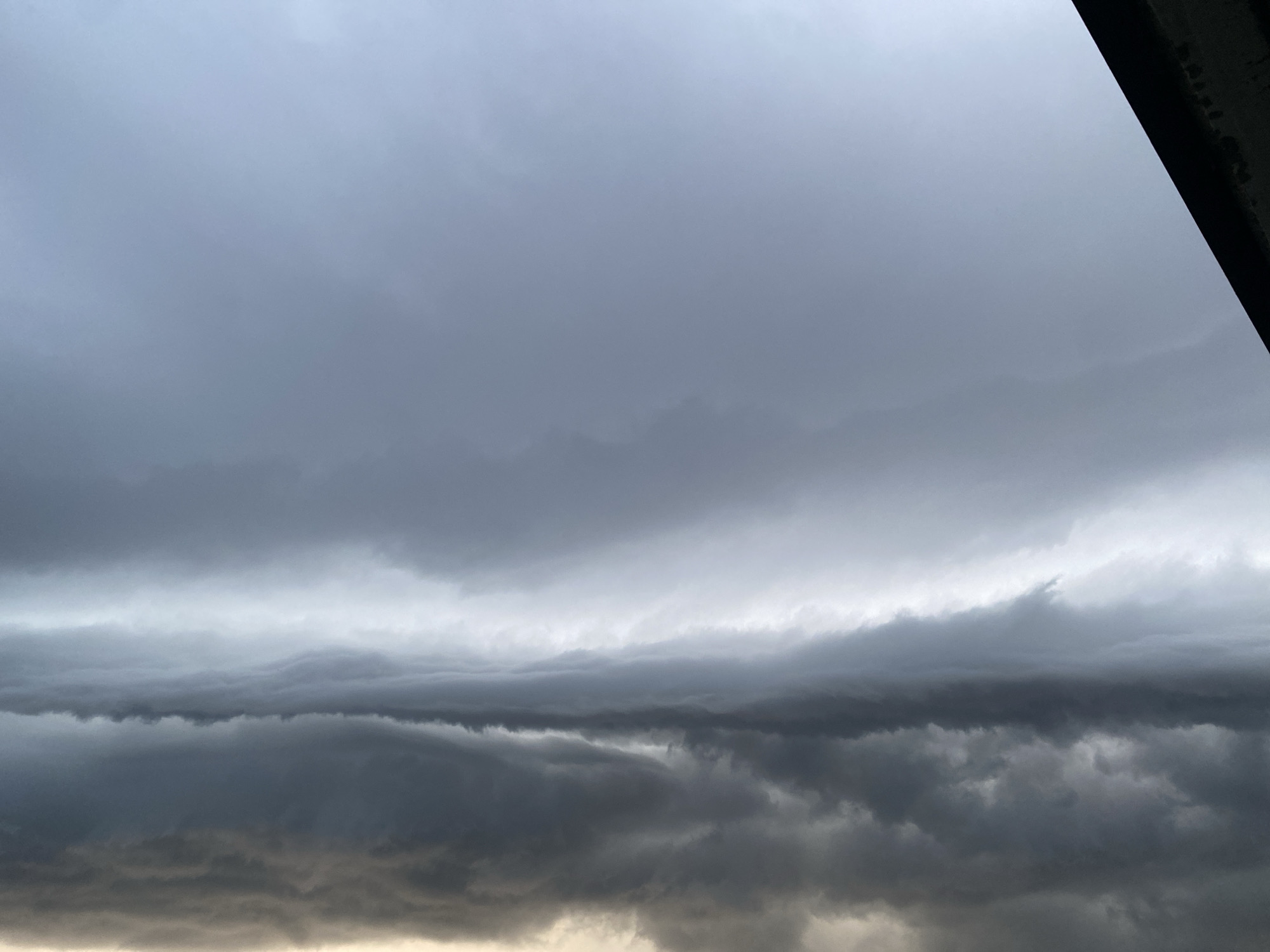 The image shows a dramatic sky filled with layers of dark, ominous storm clouds. The clouds are dense and appear to be moving, with a textured and undulating quality. The lighting is dim, suggesting either early morning, late evening, or a heavily overcast day. The top right corner of the image is partially obscured by a dark, angular structure, possibly a building or overhang. The lower portion of the sky is lighter, with a yellowish-orange hue, hinting at a distant light source or the edge of the storm. The scene is devoid of any human presence or specific landmarks, rendering the location as Unknown, Unknown.