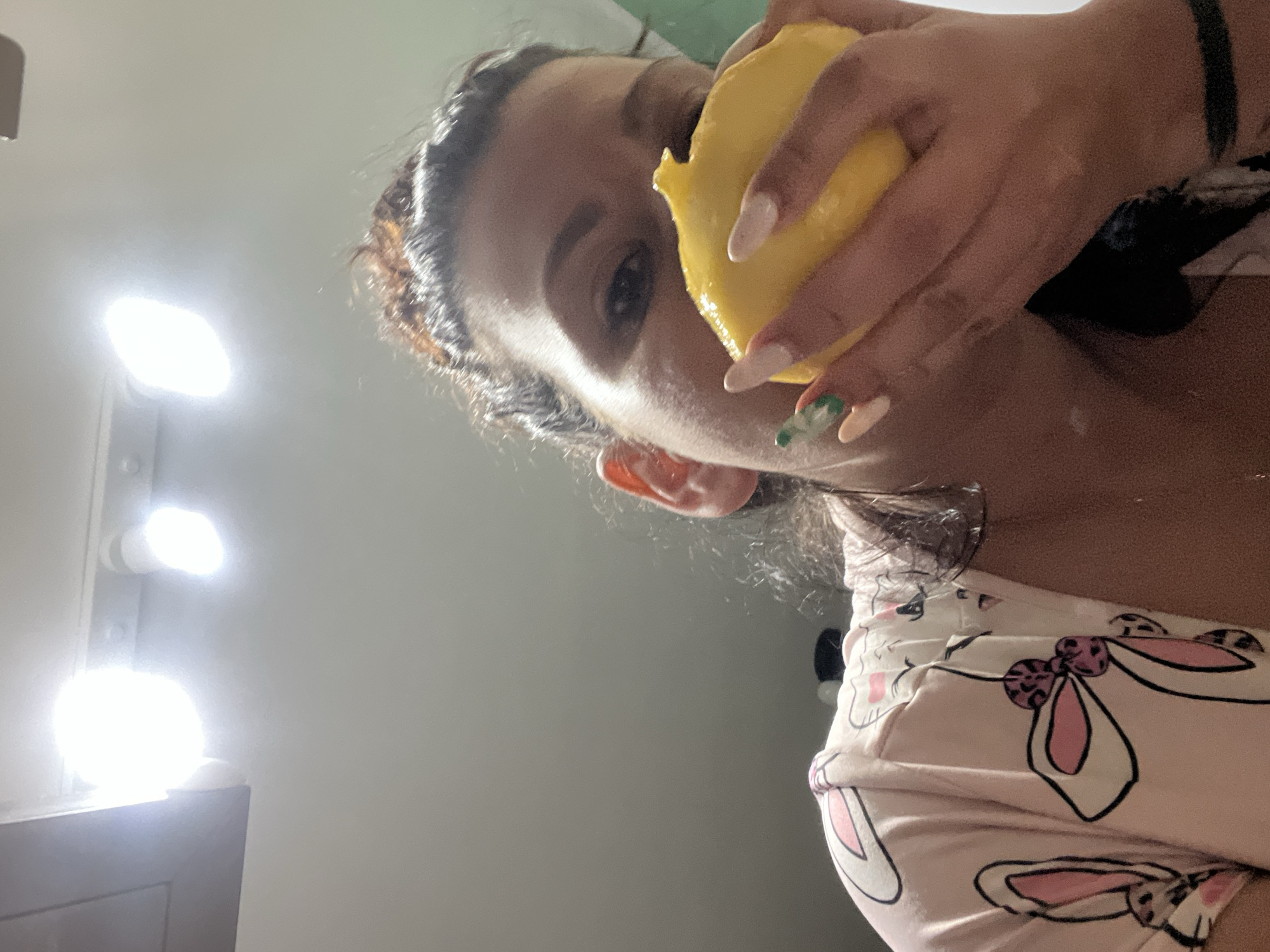 A low-angle shot depicts a person holding and appearing to bite into a yellow mango. The individual has dark hair pulled back from their face, and fair-toned skin. They are wearing a pink top patterned with white cartoon rabbit designs. Their nails are painted in light colors, with one nail adorned with a green design. The background includes a light gray wall and a vanity mirror with bright lights. The location cannot be determined as Plovdiv, Bulgaria from the image.