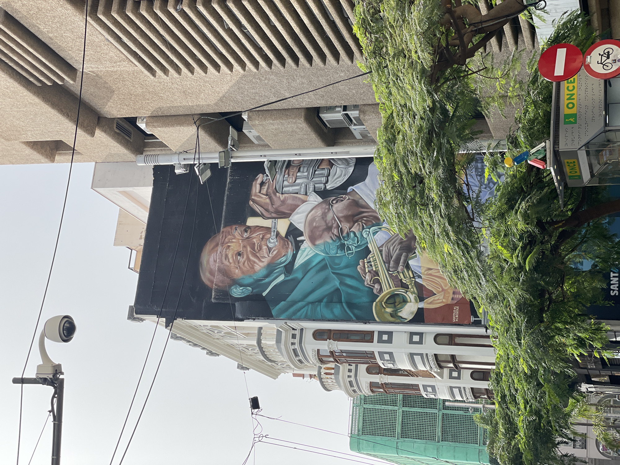 A building facade features a mural depicting a portrait of two men, one with a saxophone. Part of the mural shows medical personnel. The building is located in Santa Cruz de Tenerife, Spain. A tree partly obscures the mural. A security camera is visible on the left. Street signs and a building across the street are also visible.