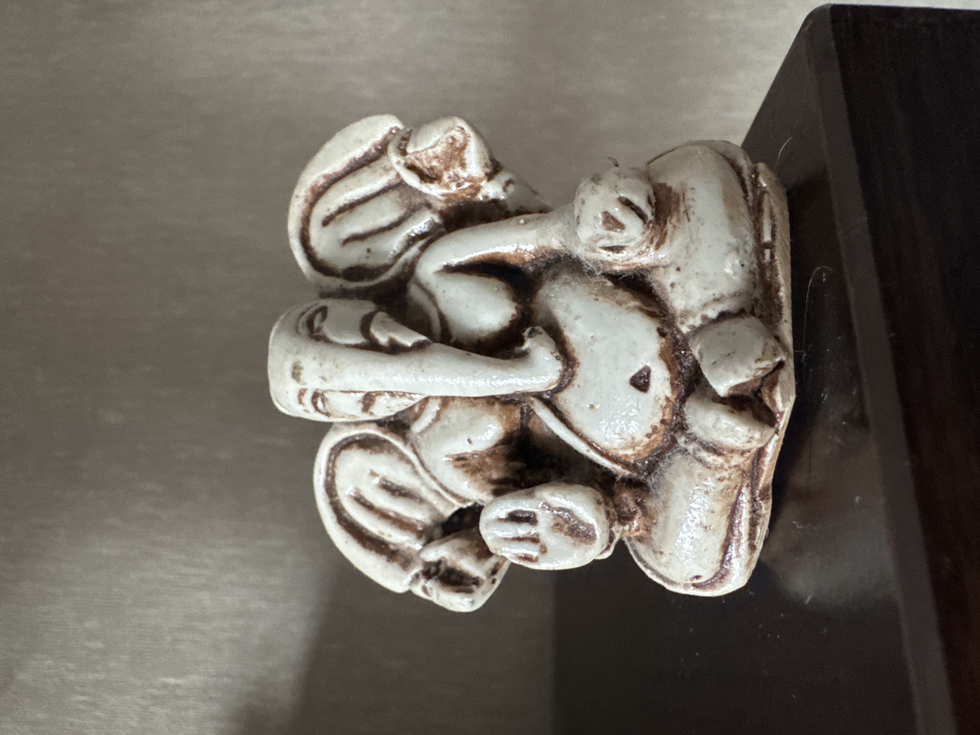 A small, antique-looking statue of the Hindu deity Ganesha sits on a dark, polished wooden surface. The statue is predominantly off-white with brown detailing that highlights the deity's features, including its elephant trunk, large ears, and seated posture. The background consists of a textured, neutral-colored wall. The image appears to be taken indoors, likely in a home or a private collection space within Bishkek City, Kyrgyzstan, though no specific landmarks or street views are visible to confirm the exact location within the city.