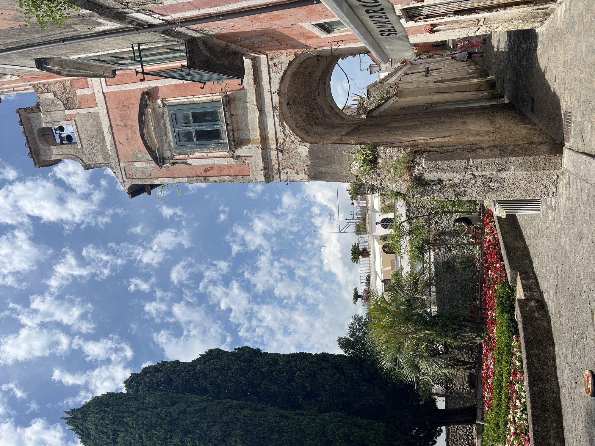 This image captures a street scene in Ravello, Italy, likely near the Duomo. In the foreground, a cobbled pathway leads into an arched passageway. To the left, a lush garden with a vibrant display of red and pink flowers is visible, bordered by a stone wall. A palm tree stands prominently in the garden, and tall cypress trees flank the scene on the left.
The archway opens to a narrow street, where two women are walking away from the camera. One woman is wearing a pink dress and a hat, and the other is in a white top and dark skirt. A sign with the text "SAMMARICO" is visible on an awning above a building on the right side of the street.
The architecture features aged, pastel-colored buildings, with one structure on the right exhibiting a weathered pink facade with a green-shuttered window and a small balcony. A belfry with two bells is situated at the top of this building, against a backdrop of a bright blue sky dotted with white clouds. The overall impression is of a historic, sun-drenched Italian town.