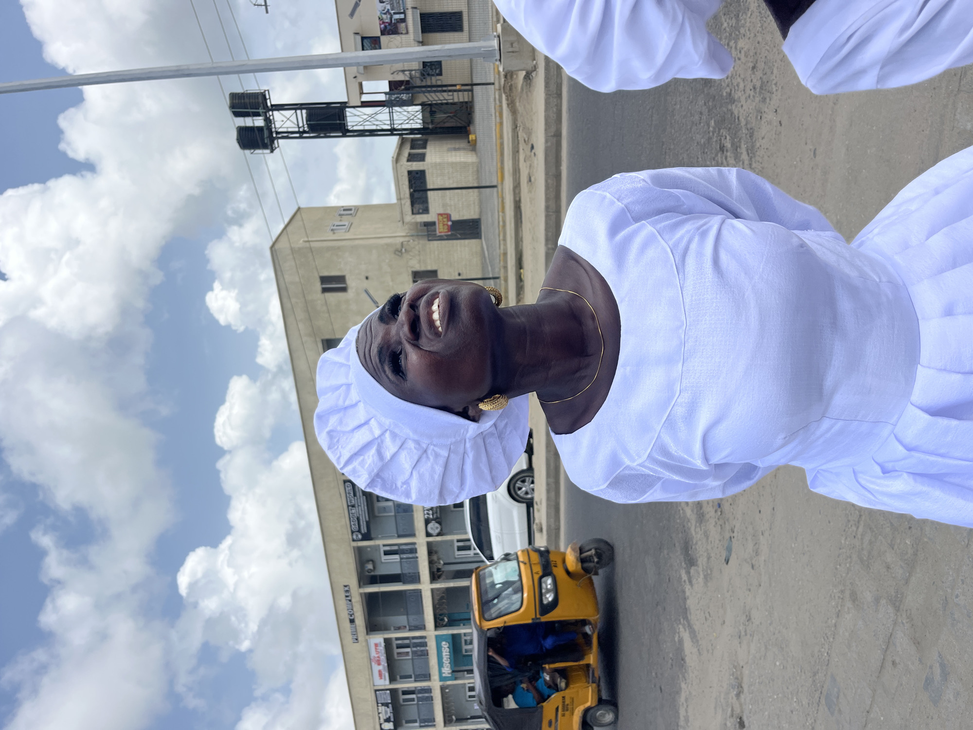 A medium-shot of a dark-skinned woman wearing a white dress and head covering in Ado, Nigeria. She is looking up and smiling broadly. She wears gold earrings and a necklace. Beside her, a person wearing white can be partially seen. A three-wheeled yellow transport vehicle sits on a gray road. The background includes buildings with signage and advertisements. The sky has fluffy white clouds. A water tower is visible behind the buildings.