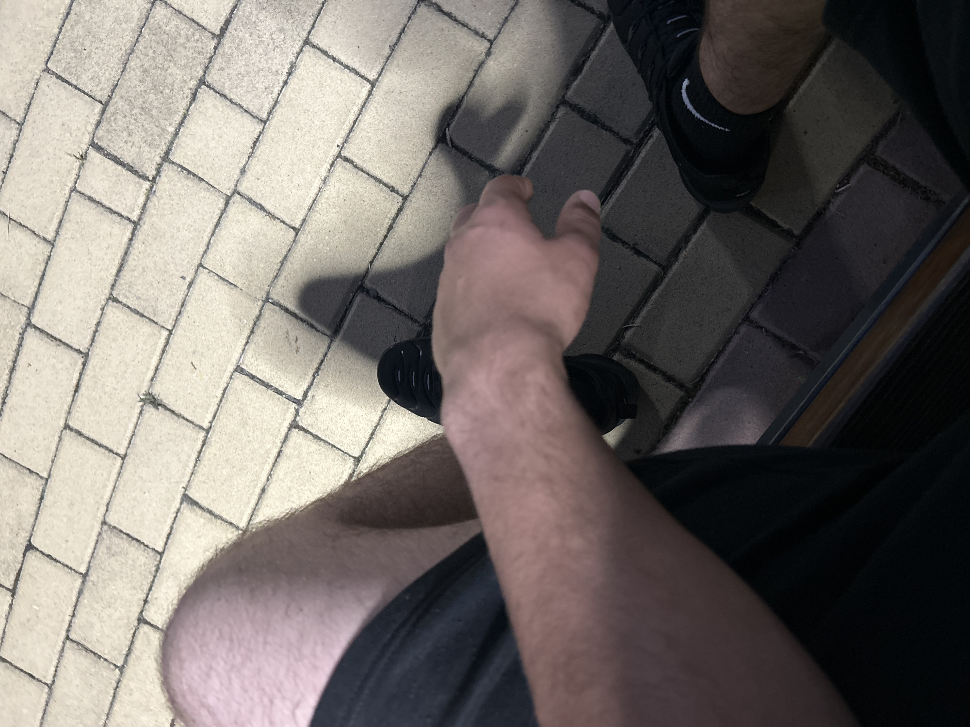 An overhead, low-light image depicts a person seated, looking down at their legs, right arm, and the ground in Dzhebel, Bulgaria. The individual's bare, hairy thighs and lower legs are visible, covered by black shorts. Black athletic shoes are worn on both feet, with a black sock featuring a white Nike swoosh visible on the right ankle. The person's bare right forearm and hand are extended forward, positioned over the paved surface. The ground consists of light-colored, rectangular paving stones laid in a staggered pattern with dark grout lines. Shadows, likely cast by the individual or an overhead light source, are visible on the paving. A dark, horizontal structure, possibly the edge of a bench or step, is present at the bottom of the frame.