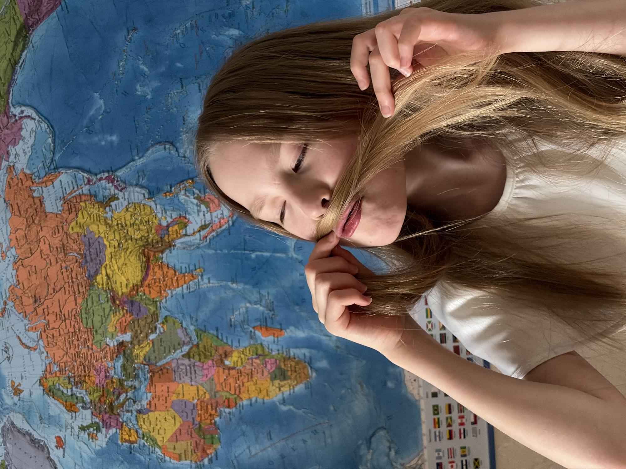 A young person with long, light brown hair, wearing a white shirt, lies on a world map. The map is multicolored, showing continents and oceans, and also displays a section with national flags. The person is holding a strand of their hair near their mouth. The location is not specified, and there is no indication it is Izhevsk, Russia.