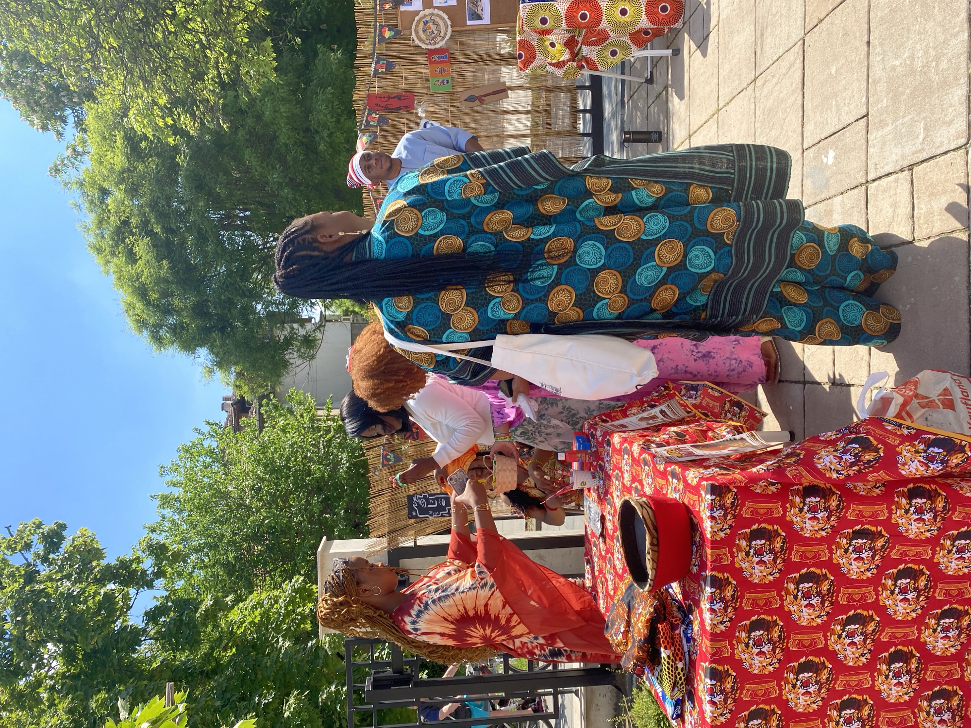 Several people are gathered at an outdoor market stall. The stall is covered with a red cloth featuring a repeating pattern of faces or masks. A woman with blonde braids is attending the stall, wearing a red and orange tie-dye top. Customers are browsing the items on display, including a woman wearing a blue and gold patterned dress with black braids, and another woman with red hair. A man in a light blue shirt and white cap is standing in the background. The market stall is set up on a paved area, with green trees visible in the background. Decorations are displayed on the back of the stall. A "Kaufland" bag is visible. The location is unconfirmed as the city of Pleven, Bulgaria.