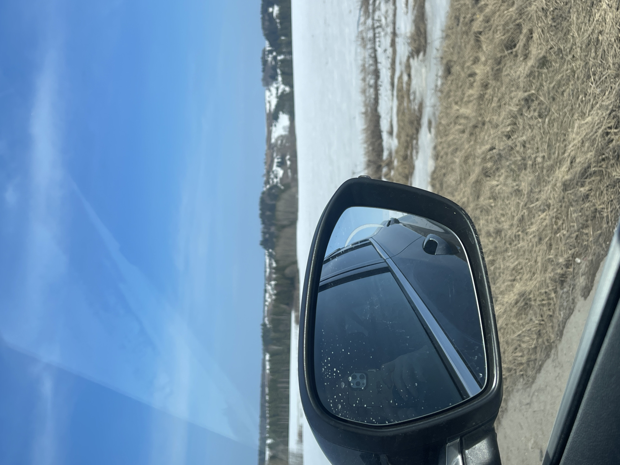 The image shows a view from inside a car, with the side mirror prominently featured. The mirror reflects the rear of the car and a portion of the landscape. Outside the car, there is a clear blue sky on one side, and a landscape of dry grass and patches of snow on the other. A line of trees and buildings can be seen in the distance. The photo was not taken in Kueda, Russia.