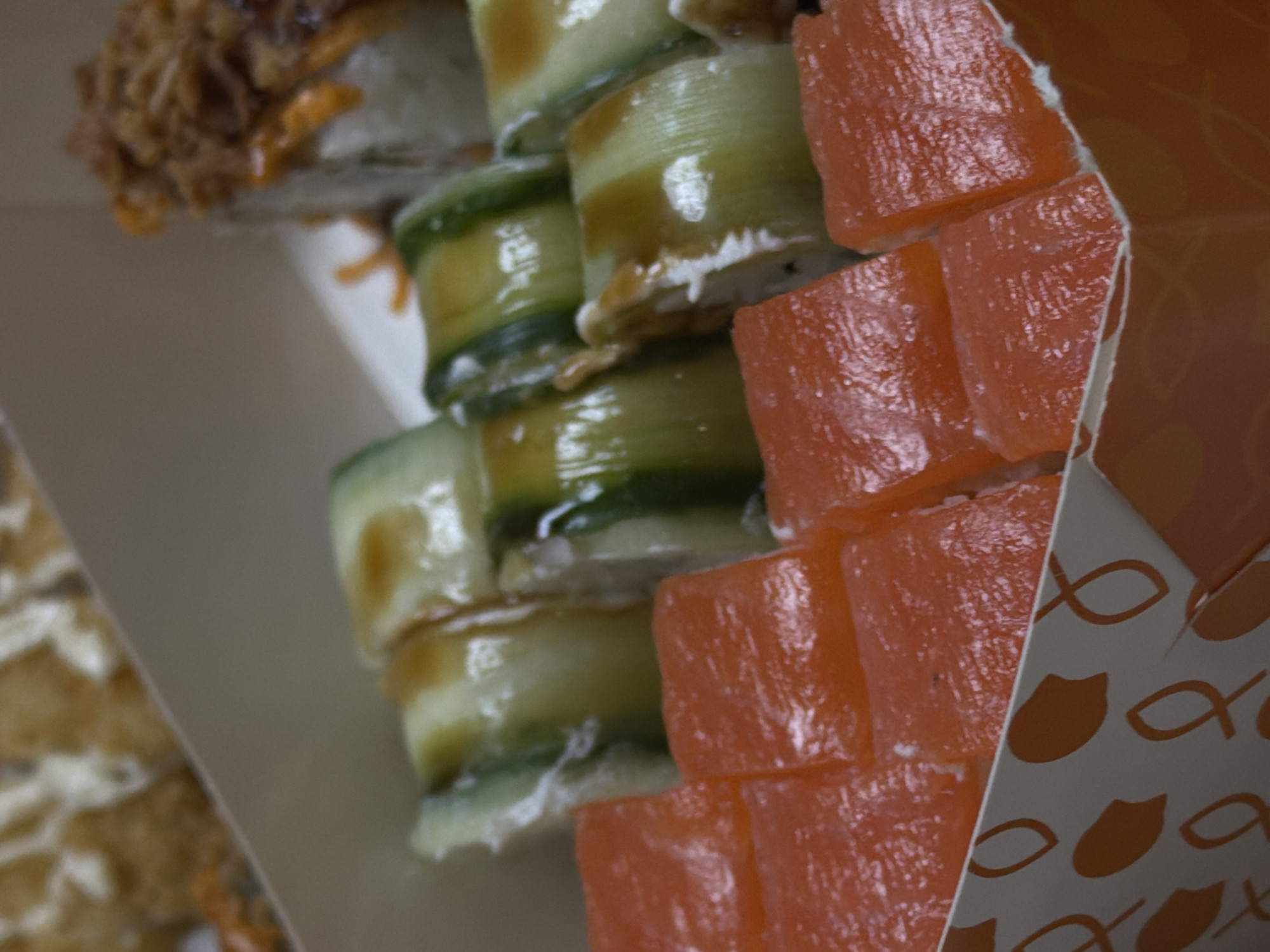 A close-up, high-angle photograph captures a selection of sushi rolls presented in what appears to be a takeout container or on a serving platter. The image features a shallow depth of field, with the food in the mid-ground sharply focused, while the foreground and background are softly blurred.
On the top left, a sushi roll is visible, adorned with a topping of what appears to be crispy fried onions or a similar golden-brown, textured ingredient, complemented by a creamy white sauce over the rice. Below and extending towards the center, several sushi rolls are neatly wrapped in thin, translucent slices of a green vegetable, likely cucumber, with white filling peeking out from the ends. To the right side of the frame, a prominent stack of vibrant orange, rectangular pieces of fish, possibly salmon, is arranged, showcasing their rich color and smooth texture.
A diagonal white line, likely the edge of a plate or container, divides parts of the composition. On the far right, a segment of a cardboard container is visible, featuring a repeating pattern of stylized orange fish shapes, suggesting packaging from a seafood-oriented establishment. No people or specific activities are depicted. The lighting is soft and diffused, highlighting the intricate details and fresh appearance of the sushi. Given the context, this dish is likely from Perm, Russia.