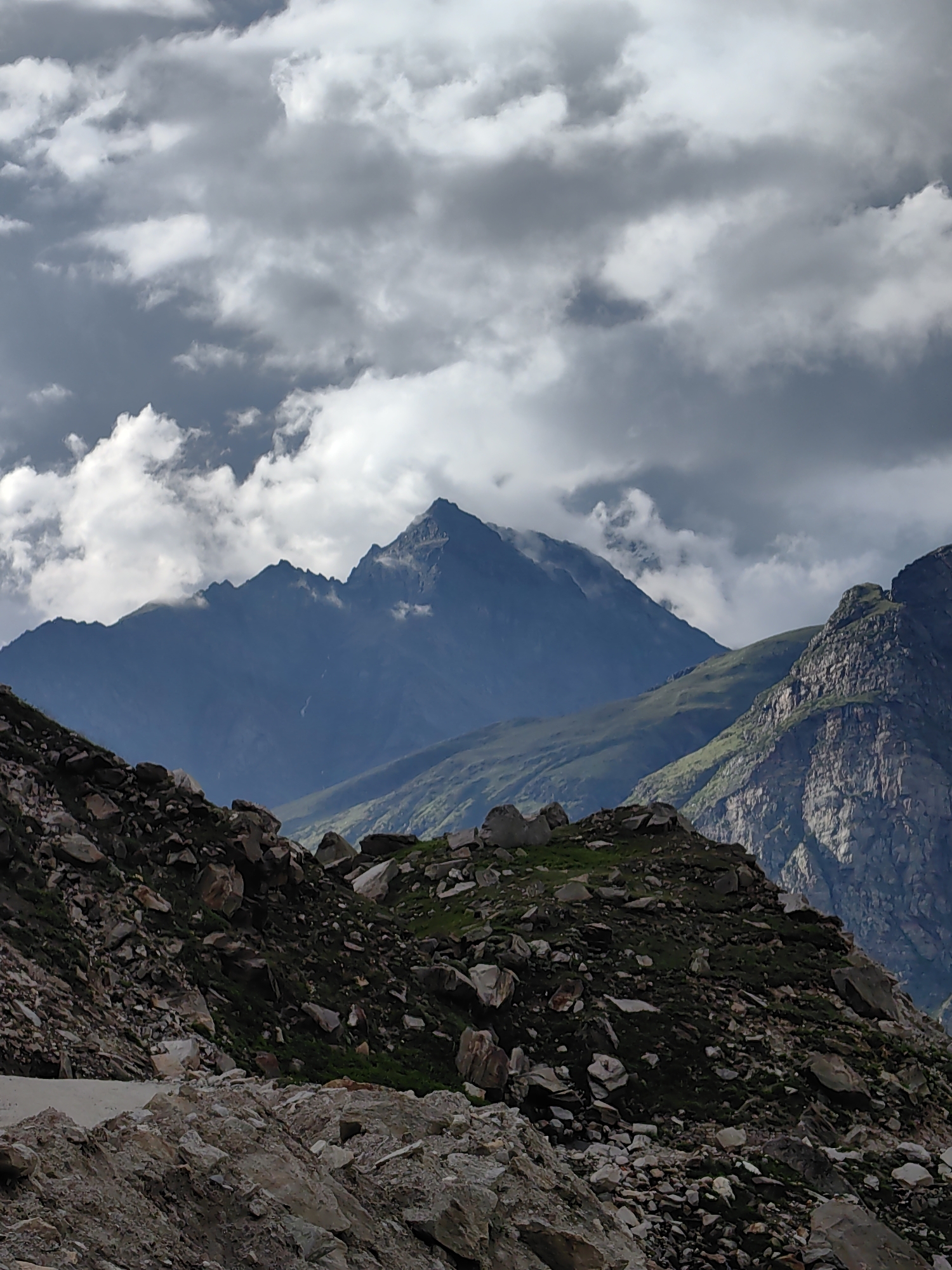 The image depicts a dramatic mountainous landscape under a cloudy sky. In the foreground, a rocky, debris-strewn slope with patches of green vegetation dominates the lower part of the frame. Further back, layers of mountains recede into the distance, their peaks sharp and imposing. The middle ground features a prominent, dark blue mountain with a triangular summit, partially obscured by swirling clouds. To the right, another rugged mountain with textured rock faces rises. The sky is filled with a mix of dark, heavy storm clouds and lighter, wispy white clouds, creating a sense of depth and atmosphere. The overall impression is one of a wild, untamed natural environment. This scene is located in the vicinity of Burwa, India, a region known for its high-altitude terrain and challenging weather conditions. There are no people or specific man-made structures visible in the image.