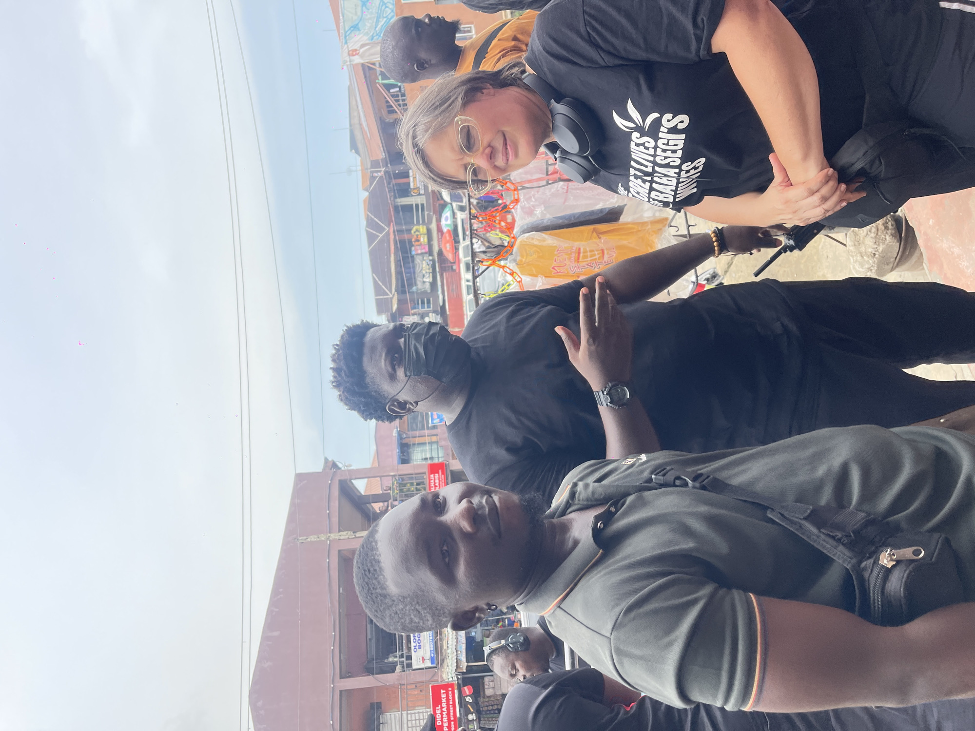 This is a daytime outdoor image featuring four individuals in what appears to be a bustling market or street setting. The image is rotated 90 degrees clockwise from its natural orientation.
In the foreground, a man with dark skin, a short beard, and a dark green polo shirt, wearing a black shoulder bag, looks towards the camera. Behind him and slightly to his right, another man with dark skin wears a black t-shirt and a black face mask, making a "V" or peace sign gesture with his right hand, revealing a wristwatch.
To the right of the frame, a woman with fair skin, glasses, and headphones around her neck smiles faintly. She wears a black t-shirt with white text that reads "GRET LIVES BABA SEGI'S WIVES". Partially visible behind her is another man with dark skin in an orange or brown shirt.
The background shows multiple multi-story buildings, likely shops or market stalls, with various goods and structures visible. Bright blue power lines stretch across the clear, bright sky. Visible text on signs includes "DIDEL SUPERMARKET", "ON STREET BLOCK 3", "ALHAJA LABISI", and "OLOR BOG", suggesting a commercial area. The overall scene conveys a lively, open-air environment.