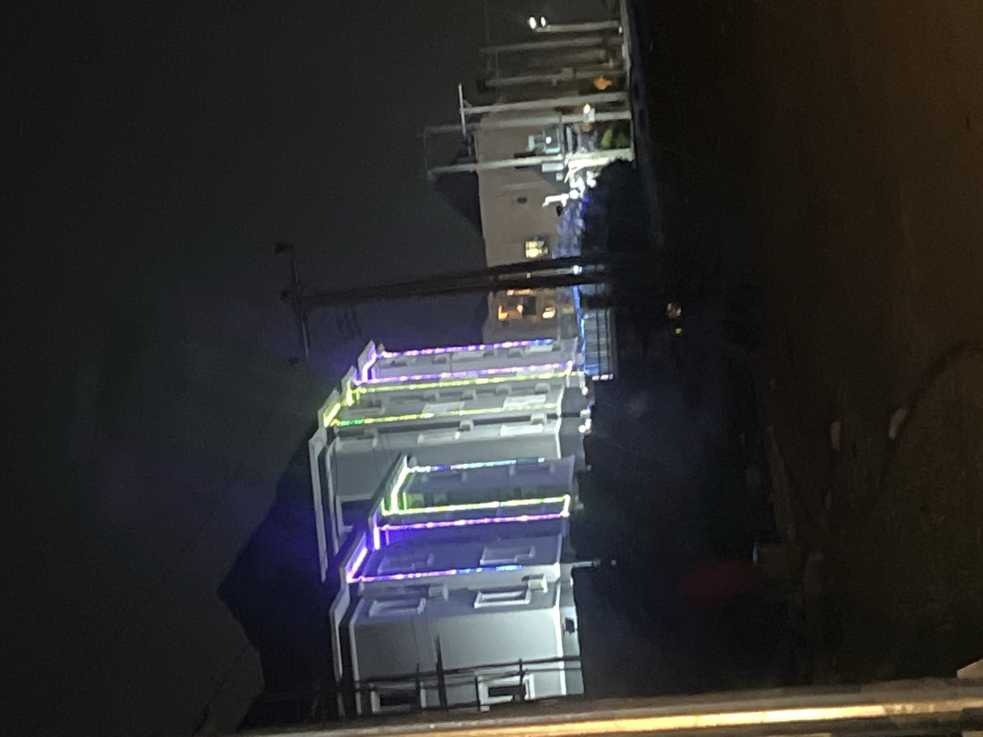 The image, captured at night and rotated vertically, depicts a scene from Gwarinpa, Nigeria, showcasing several multi-story buildings adorned with vibrant, colorful lighting. The primary subjects are these modern-looking buildings, each featuring multiple levels and outlined with decorative LED strip lights.
The lighting display is a notable feature, with distinct lines of purple, blue, green, and yellow lights tracing the facades of the buildings, likely along balconies or architectural elements. Some windows within the buildings show internal illumination, indicating occupancy. In the background, other structures are faintly visible, and a utility pole or similar tall, dark structure stands to the left. The sky is dark, typical of a nighttime setting, providing a strong contrast to the brightly lit buildings.
No people or specific activities are discernible, presenting a static urban or suburban nightscape. The foreground includes dark, blurry elements, possibly a railing or an obstruction from where the photo was taken. There is no visible text in the image.