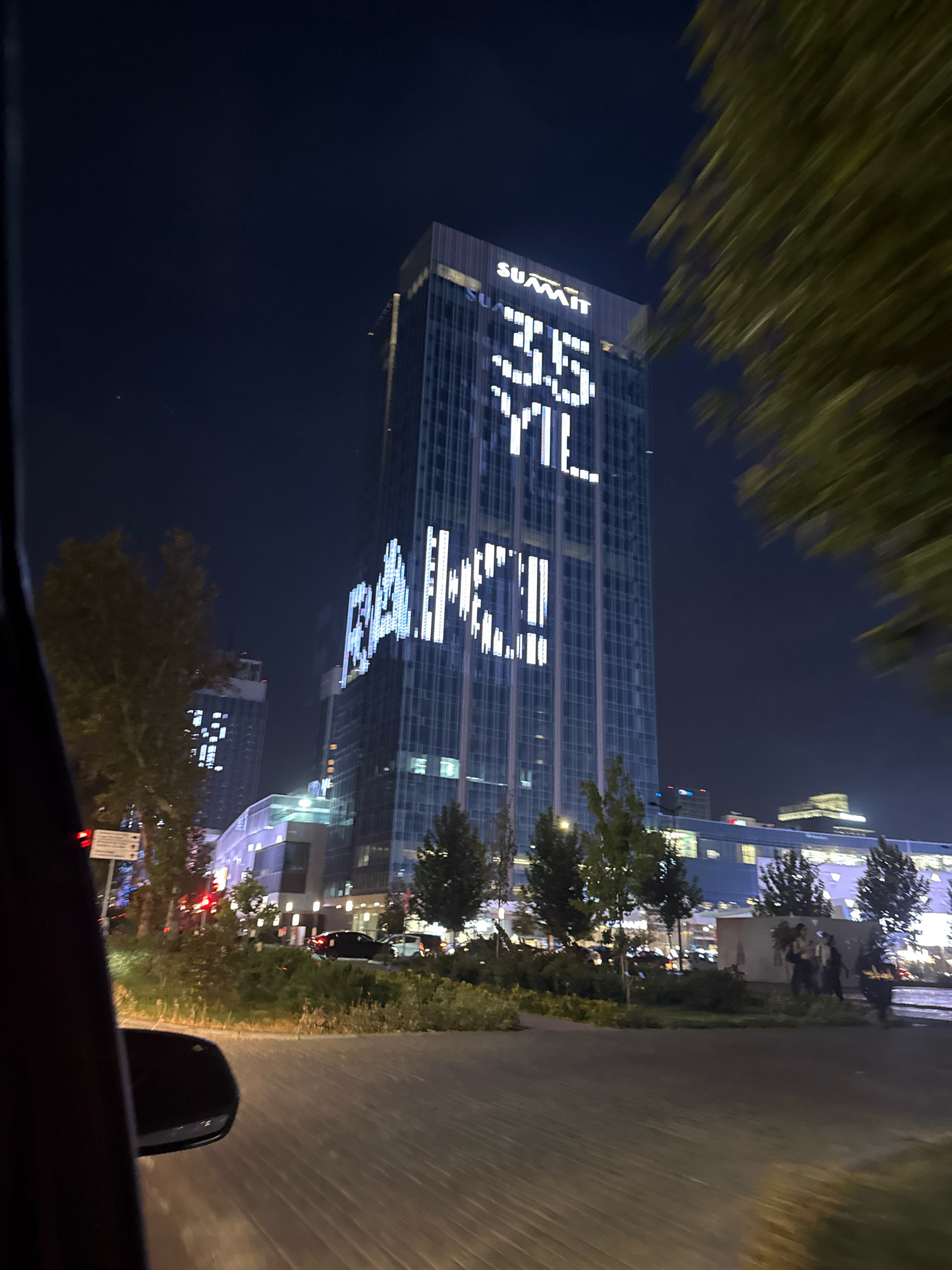 This is a nighttime, exterior, eye-level, medium shot captured from a vehicle, looking towards a tall, modern skyscraper in Tashkent, Uzbekistan. The skyscraper, likely part of a complex, displays illuminated text on its facade. At the top, the word "SUMMIT" is visible in white capital letters. Below that, large white numerals "35" are prominently displayed, followed by "YL" in a similar font, suggesting a 35th-anniversary celebration. Further down the building, larger, more abstract illuminated text spells out "BAKCI" in white. The skyscraper is surrounded by other buildings, some with illuminated windows, and a low-lying structure with a patterned facade. Trees with sparse foliage are scattered throughout the foreground and middle ground. The street in the foreground is paved, and traffic lights are visible in the distance. The overall atmosphere is that of a modern, urban environment at night.