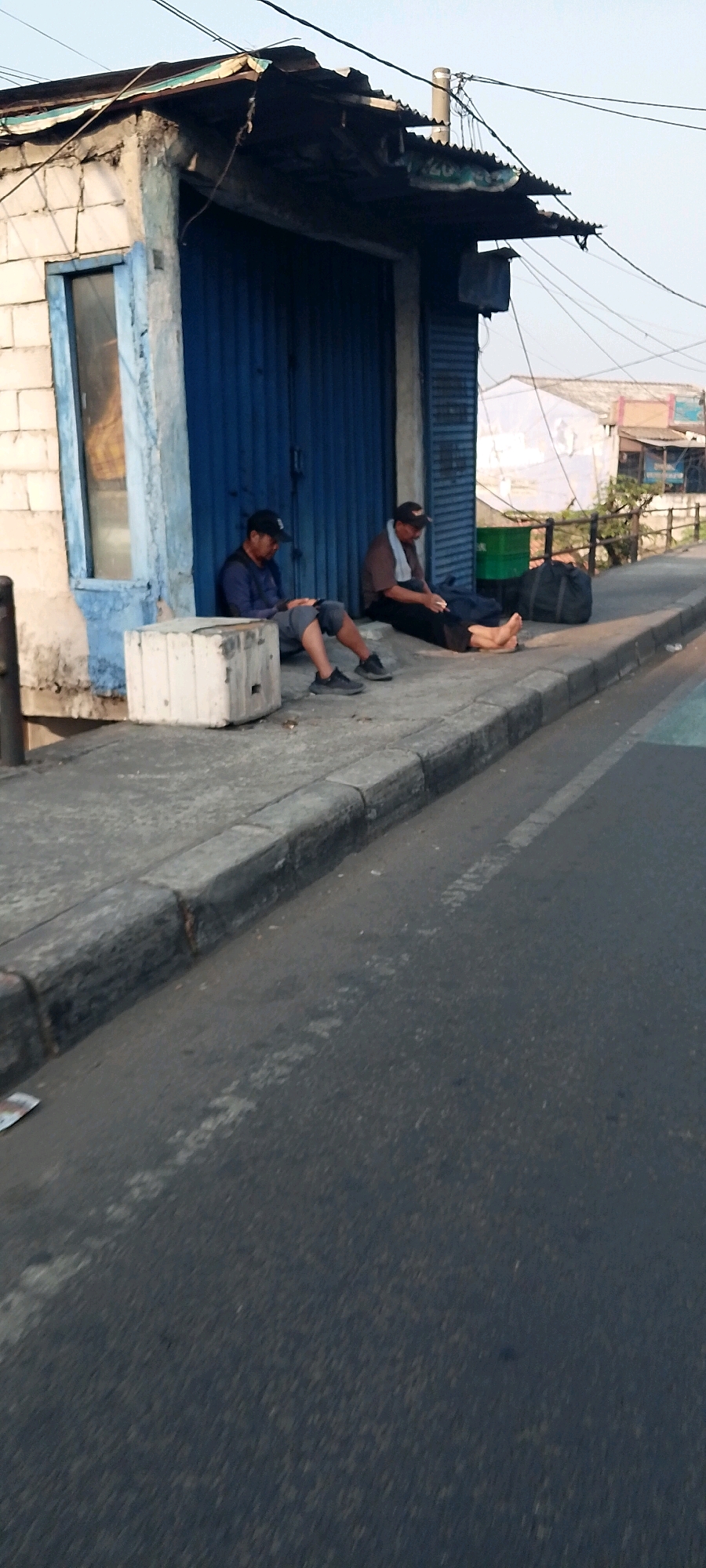 Two men are sitting on a sidewalk in front of a blue shuttered building. The building appears to be a shop or stall, with a corrugated metal roof and a concrete block structure. One man is wearing a dark blue long-sleeved shirt and a black baseball cap, and is looking down at his phone. The other man is wearing a brown t-shirt, a towel around his neck, and is also looking down, possibly at a phone or cigarette. A large black bag and a green plastic bin are visible behind the men. The sidewalk is made of concrete and there is a curb separating it from a street with a white line. In the background, there are other buildings and some vegetation. The scene suggests a casual or resting moment in an urban environment in Bekasi, Indonesia.