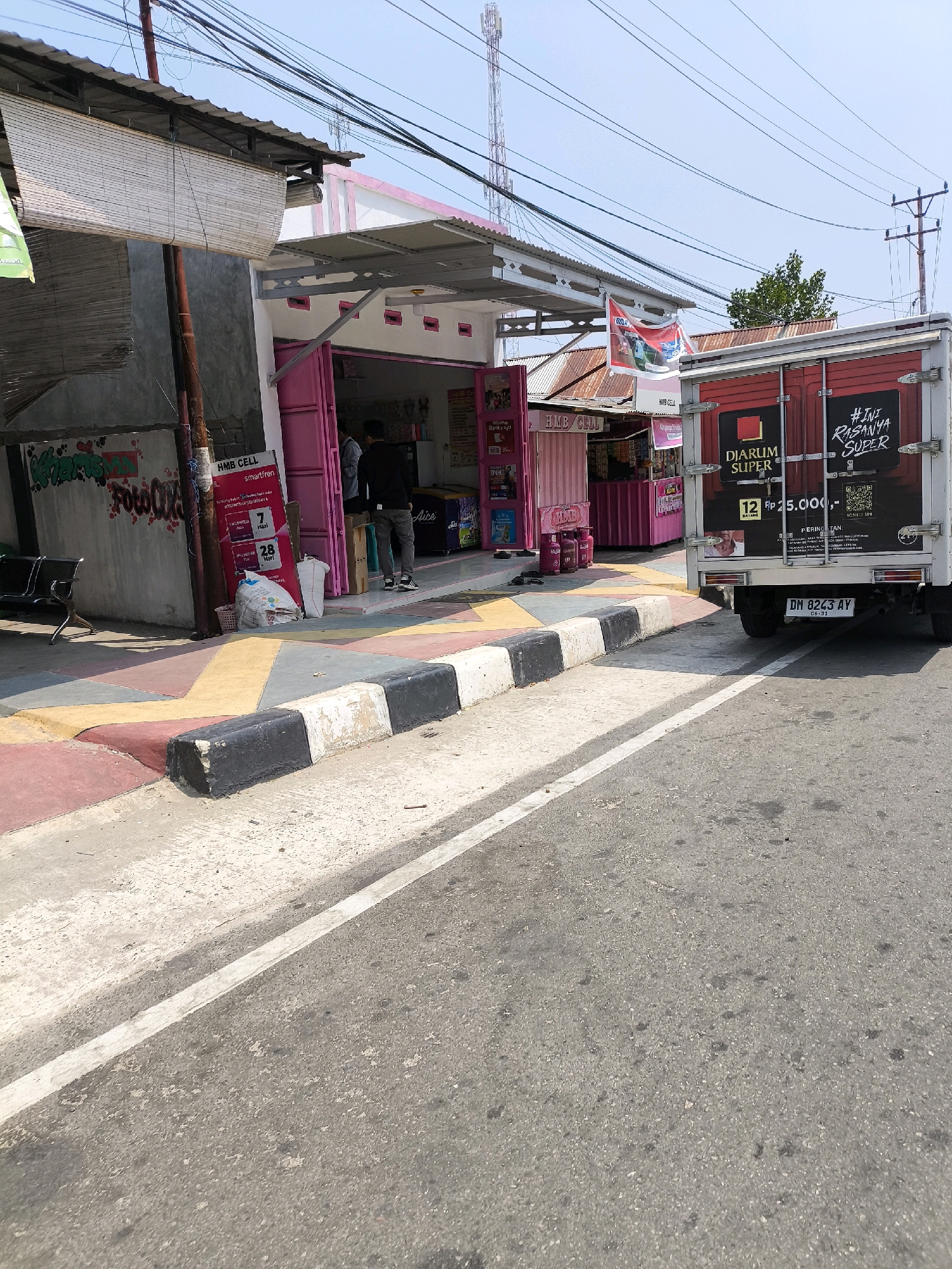 The image captures a street scene in Kwandang, Indonesia, on a bright, sunny day. In the foreground, a section of asphalt road is visible, marked with a white line. A raised concrete curb, painted with alternating black and white blocks, separates the road from a sidewalk. The sidewalk itself is adorned with colorful geometric patterns in yellow, pink, and gray.
To the left of the sidewalk, a building with a grey concrete wall is partially visible, covered with graffiti. A bamboo blind shades a portion of this building. Adjacent to it, a pink and white storefront, identified as "HMB CELL," is open for business. Several pink gas canisters are placed outside the store. Inside, a few individuals are present, including one person in a black shirt and grey pants standing near the entrance, and another further inside.
A red delivery truck with "DIARUM SUPER" branding is parked on the right side of the street, partially obscuring the view of another building behind it. The truck features graphics and text, including what appears to be promotional material. A license plate reading "DM 8243 AY" is visible on the rear of the truck. In the background, telephone poles with numerous wires crisscross the sky, and a tall communication tower stands prominently against the clear blue sky. Trees are also visible in the distance. The overall impression is of a typical commercial street in a developing urban area.