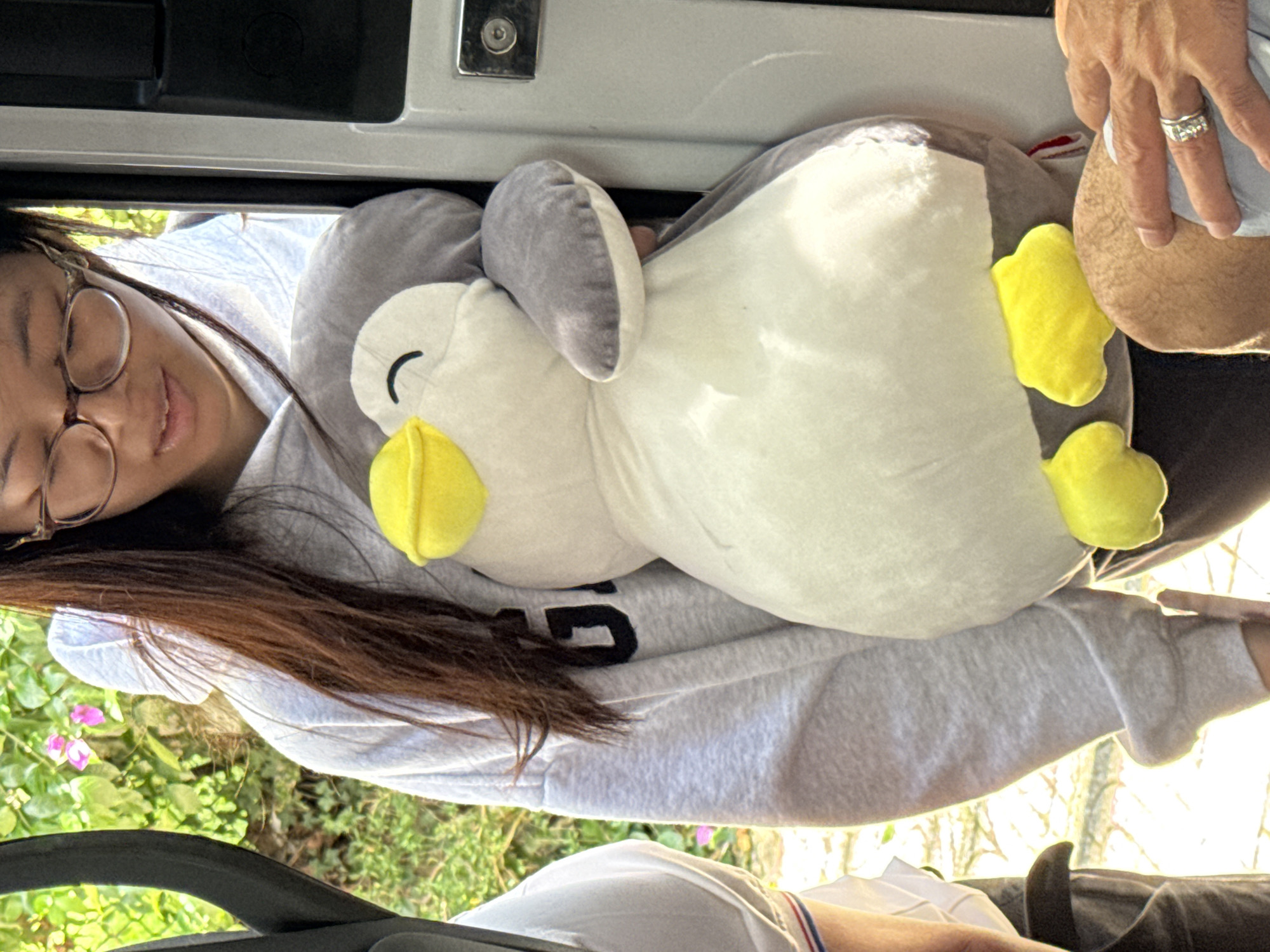 In Phường Xuân Hương - Đà Lạt, Vietnam, a young woman with long brown hair and glasses is holding a large stuffed penguin. She is wearing a gray hoodie with black lettering on it. The penguin is white and gray with bright yellow feet and a yellow beak. The woman is standing next to an open white door or vehicle. In the background, there is lush green foliage and some pink flowers, suggesting an outdoor setting. Another person's hand with a ring on it is visible in the lower right corner of the image, resting on what appears to be a leg. The scene has a casual and relaxed feel.