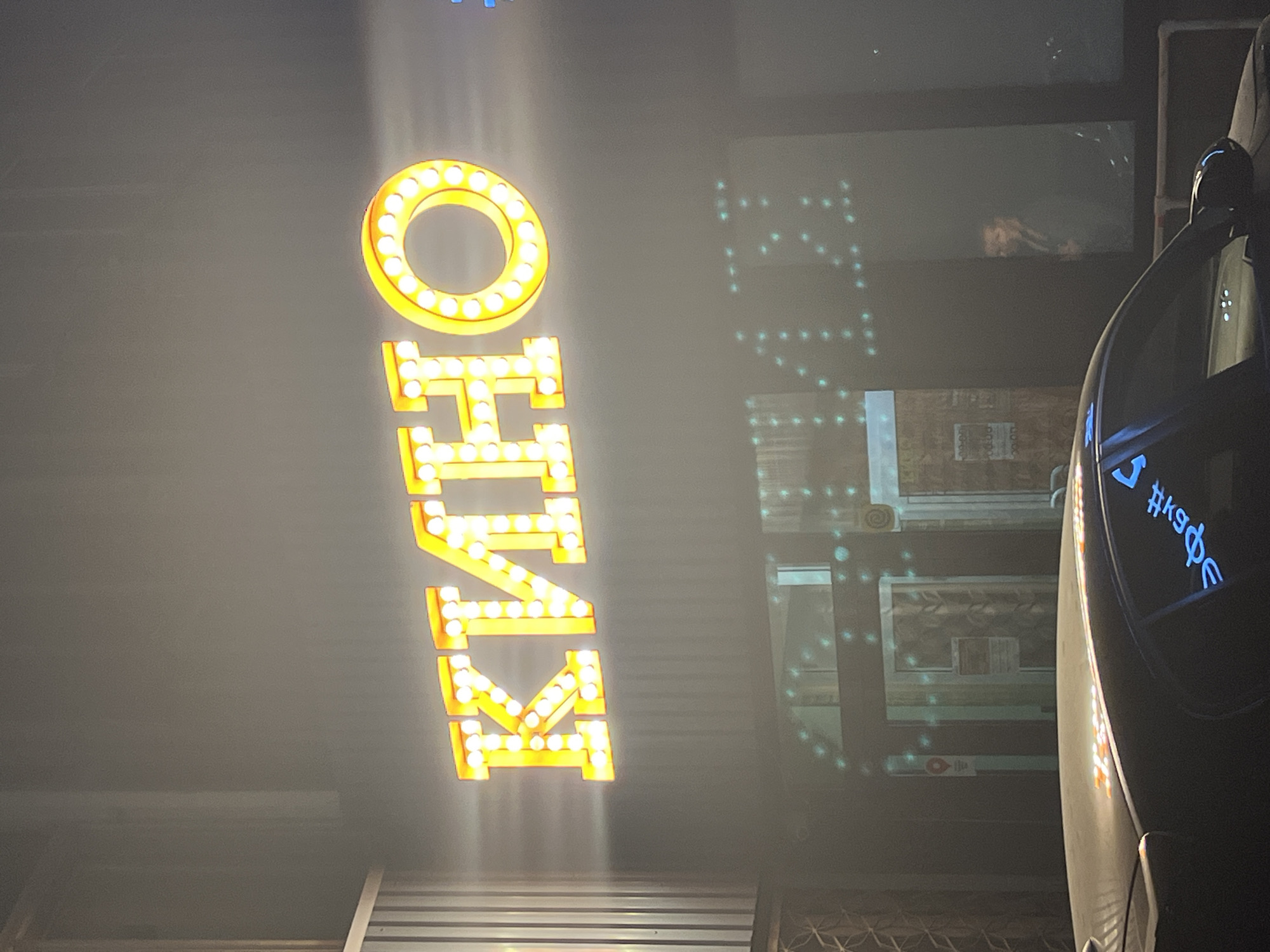 The image shows a vertical marquee sign spelling out "KINO" in large, lit yellow letters, next to a dark car. Reflected in a shop window behind the sign are indistinct images of what appear to be architectural details from a building. The sign and reflection suggest the location is Izhevsk, Russia, as "KINO" means "cinema" in Russian.