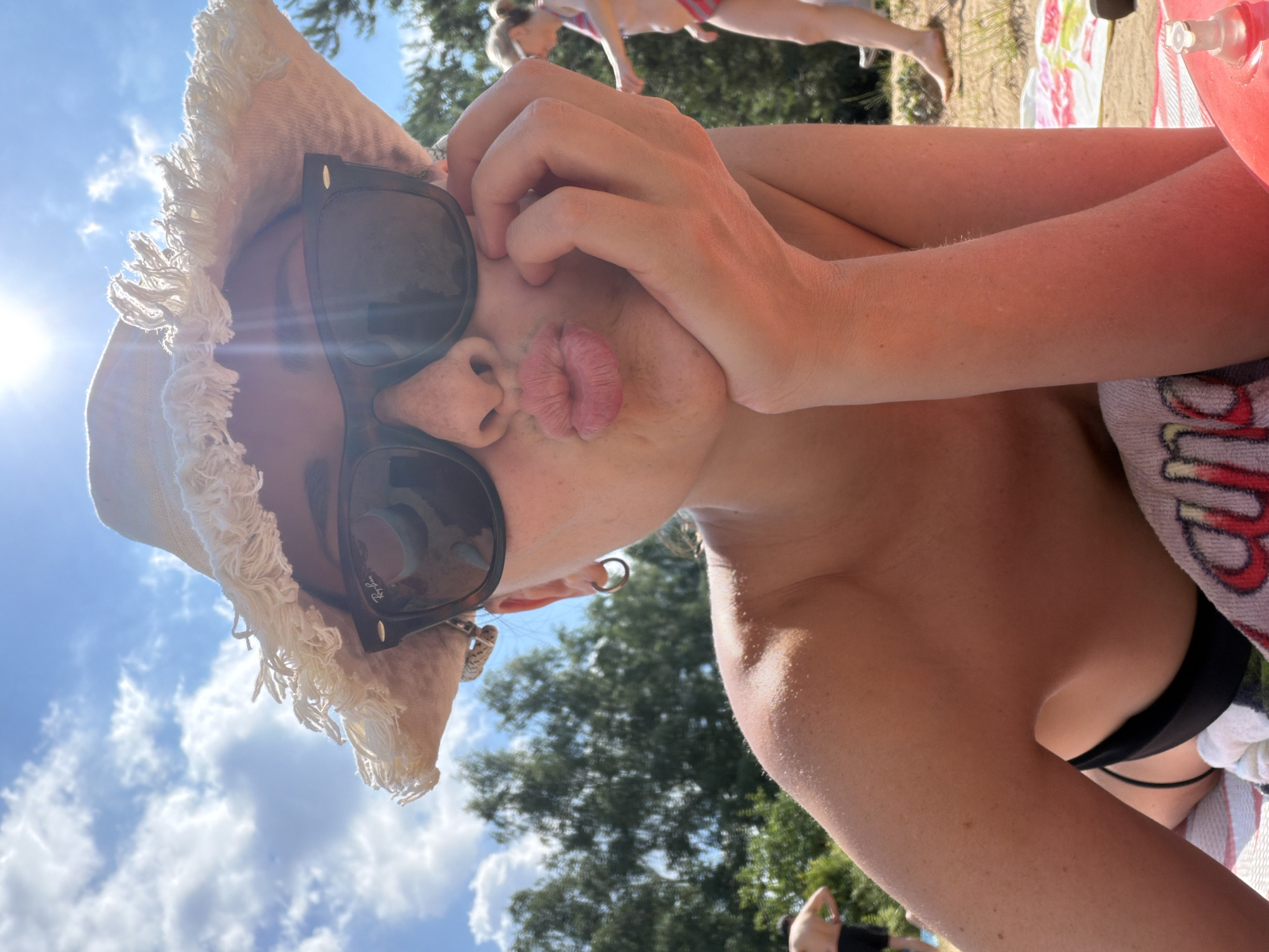 A woman is lying on a towel in Malakhovka, Russia, on a sunny day. She is wearing sunglasses and a straw hat, and she is pouting. Her hand is on her chin. In the background, other people are visible. The scene appears to be a beach or a park.