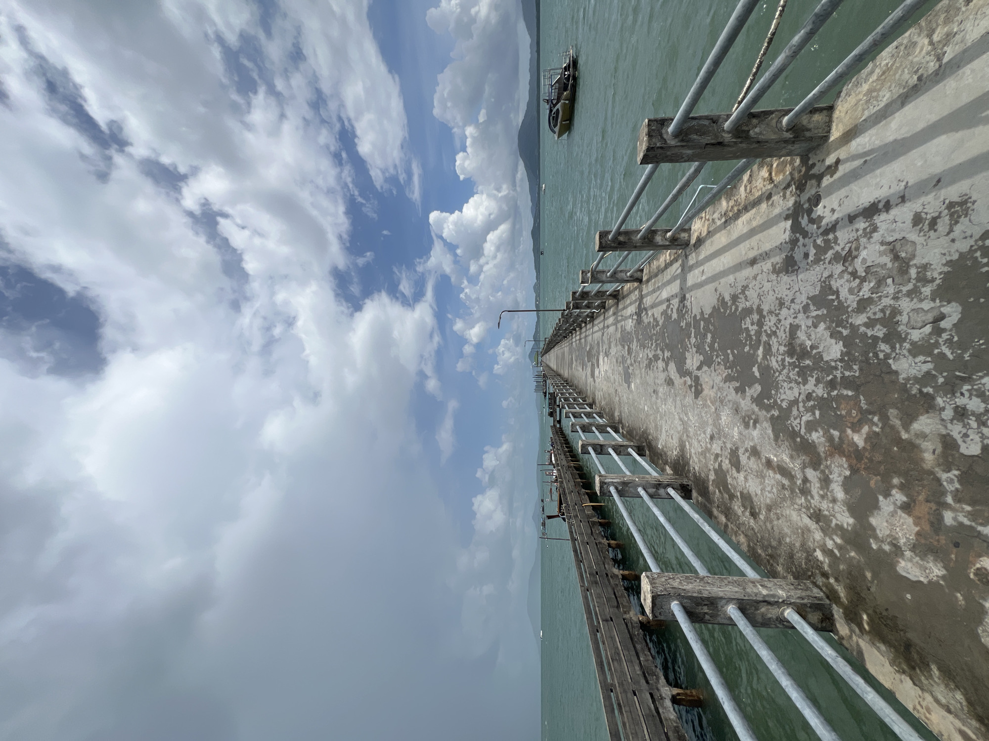 A concrete pier extends into a turquoise sea under a partly cloudy sky. The pier has a worn concrete surface and is lined with a metal railing supported by concrete posts. To the left of the concrete pier, there is an older wooden pier. On the right side of the concrete pier, a speed boat with a yellow hull is moored in the water. In the distance, a coastline with hills and some buildings is visible. The sky features dramatic white cumulus clouds interspersed with patches of blue and areas of darker, more ominous clouds on the left, suggesting possible incoming rain. The scene captures a view of the sea and the approach to the water from Pa Khlok, Thailand.
