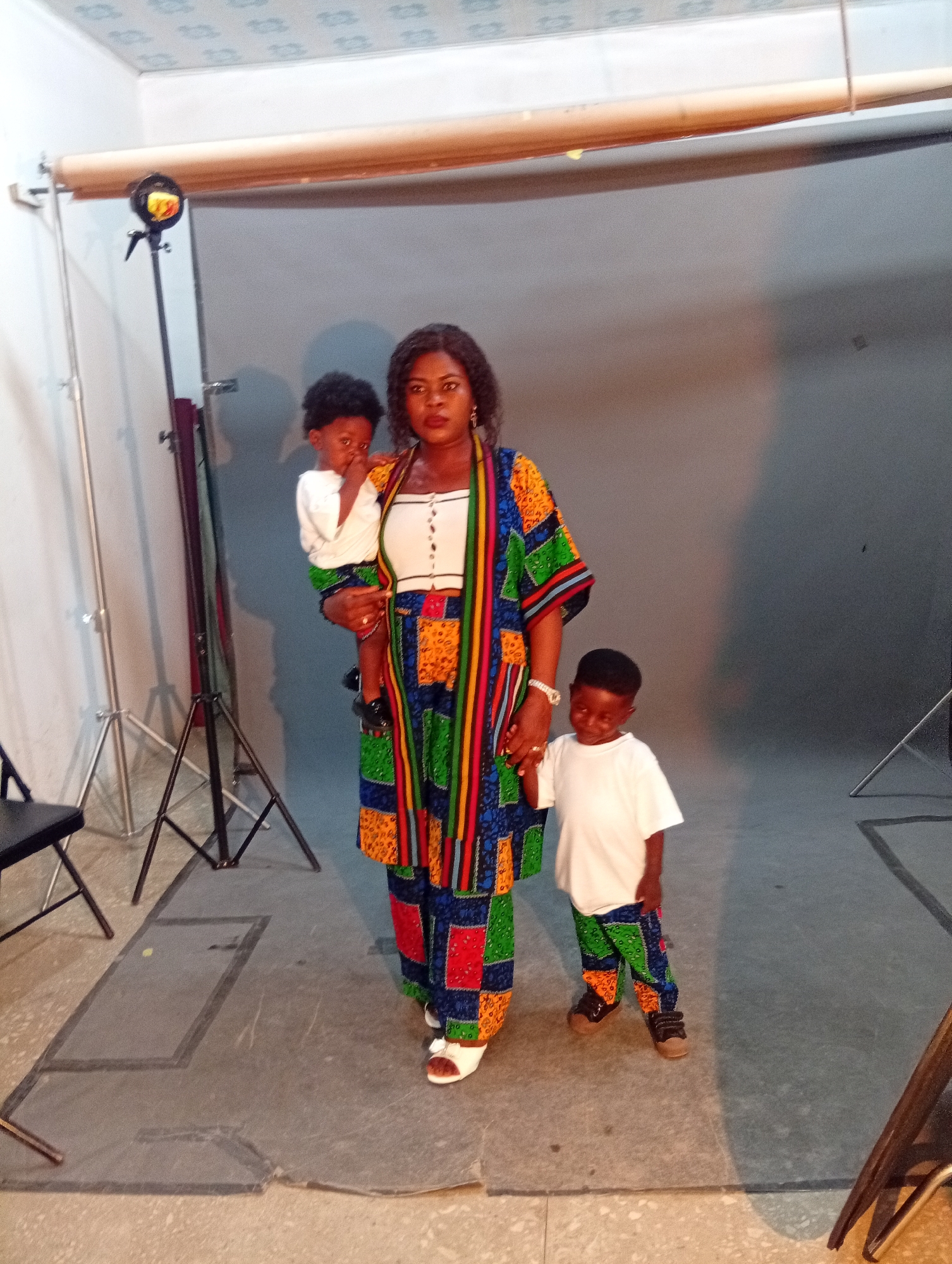 In a studio setting with a grey backdrop, a Black woman stands holding a young Black boy. She is wearing a colorful African print ensemble consisting of a patterned kimono-style top and matching pants. The woman has dark, curly hair and is looking directly at the camera with a neutral expression. The boy she is holding is wearing a white t-shirt and a patterned skirt. To the woman's right, another young Black boy stands, also wearing a white t-shirt and patterned pants. He is looking down and to his left. In the background, studio equipment, including light stands and a backdrop support, are visible. A black chair is partially visible on the left side of the image. The floor appears to be a textured grey surface.
