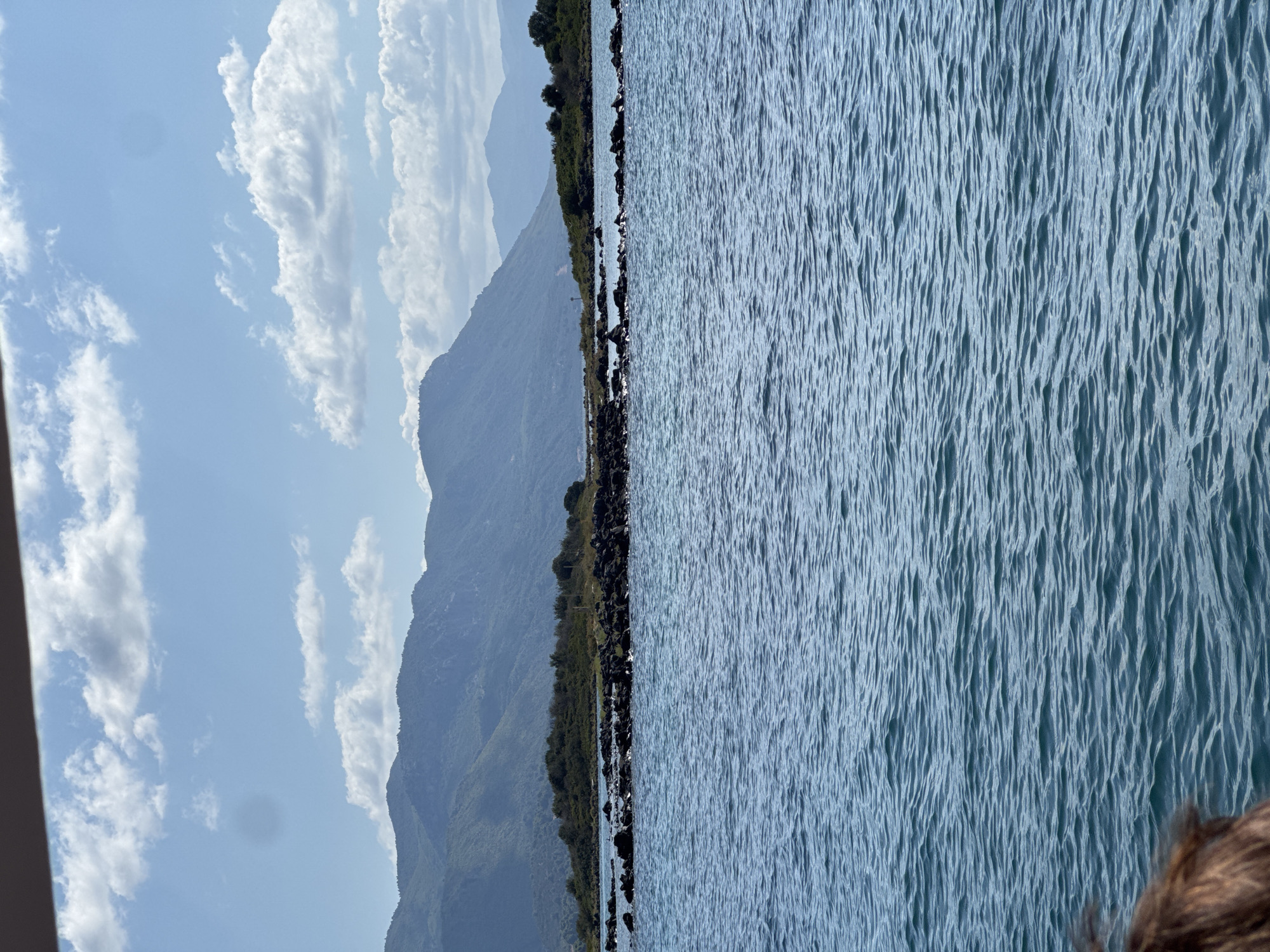 An eye-level shot captures a serene view of the Aegean Sea near the city of Unknown, Greece. The water, a vibrant turquoise, is calm with gentle ripples creating a mesmerizing pattern across the surface. In the distance, a rocky shoreline emerges, dotted with sparse green vegetation. Beyond the shore, a range of majestic mountains forms a dramatic backdrop against a clear blue sky, adorned with scattered white clouds. A dark, textured object, possibly the edge of a boat or structure, partially frames the top of the image. The overall scene is tranquil and picturesque, highlighting the natural beauty of the Greek coastline.