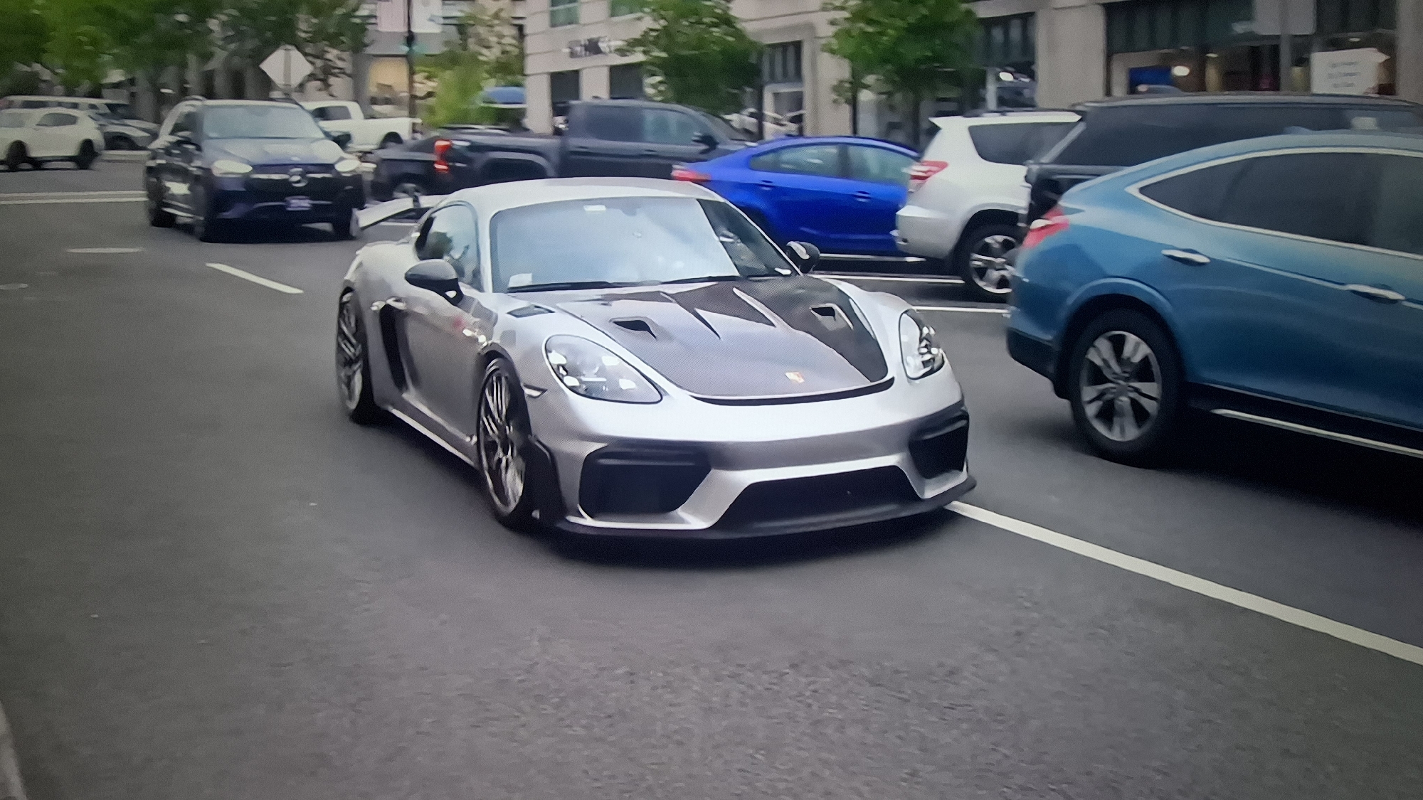 The primary subject of the image is a silver Porsche sports car, resembling a Cayman GT4, prominently featured in the foreground. It has a distinctive carbon fiber hood with integrated air vents, sleek headlights, and multi-spoke dark wheels. The car's front bumper includes large, aggressive air intakes.
The scene is an urban street, likely during the daytime under diffused or overcast lighting conditions, as indicated by the lack of strong shadows. The Porsche appears to be in motion, positioned within a lane marked by white lines on the asphalt road.
Several other vehicles populate the street around the Porsche, indicating active traffic. To the right of the Porsche is a blue Honda Crosstour or a similar compact crossover, distinguishable by its body style and unique wheel design. Further back and to the left, a dark-colored Mercedes-Benz SUV or sedan is visible. Other vehicles in the background include a white SUV (possibly a Toyota 4Runner) and a dark pickup truck or larger SUV. Buildings with multiple stories and some foliage from trees line the street in the background, suggesting a city environment.
No specific human subjects are clearly visible, although the presence of multiple cars implies drivers. There is no legible text on any of the vehicles, signs, or buildings within the image. The Porsche emblem is on the hood but is not readable. The overall impression is a bustling city street scene with a focus on high-performance automobiles.