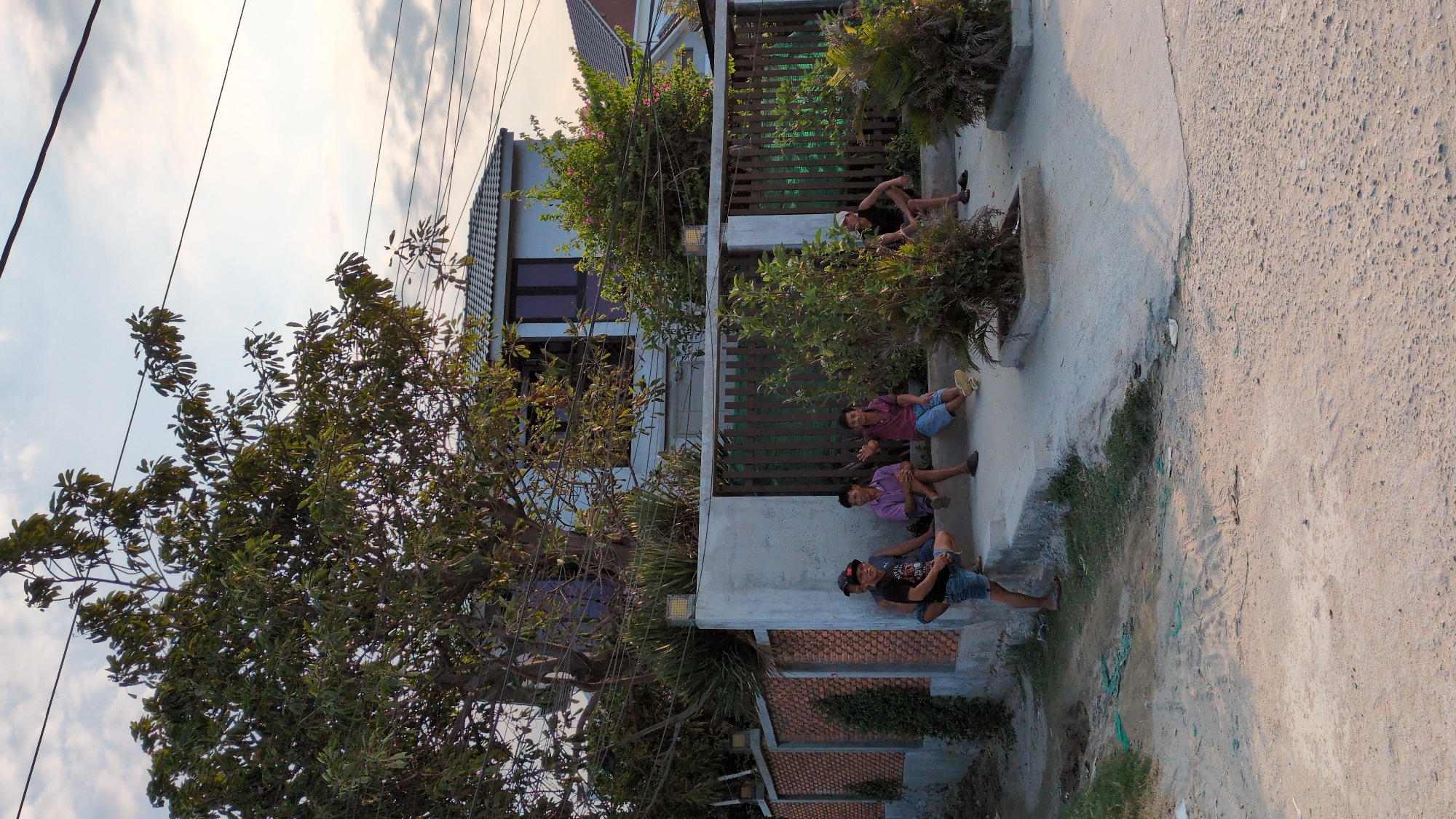 In Phan Rang – Tháp Chàm, Vietnam, several individuals are seated in front of a property next to a concrete path. One person, wearing a black shirt and hat, is seated closer to the camera with their legs extended. Two others are seated nearby, one in a purple shirt and blue shorts, and another in a maroon shirt and blue shorts. Further back near a fence, another person is seen lying down. The property is enclosed by a green fence with plants growing around it. A large tree is present on the left side of the image, obscuring part of the building. Overhead power lines are visible in the sky. The ground next to the path has scattered grass and debris.
