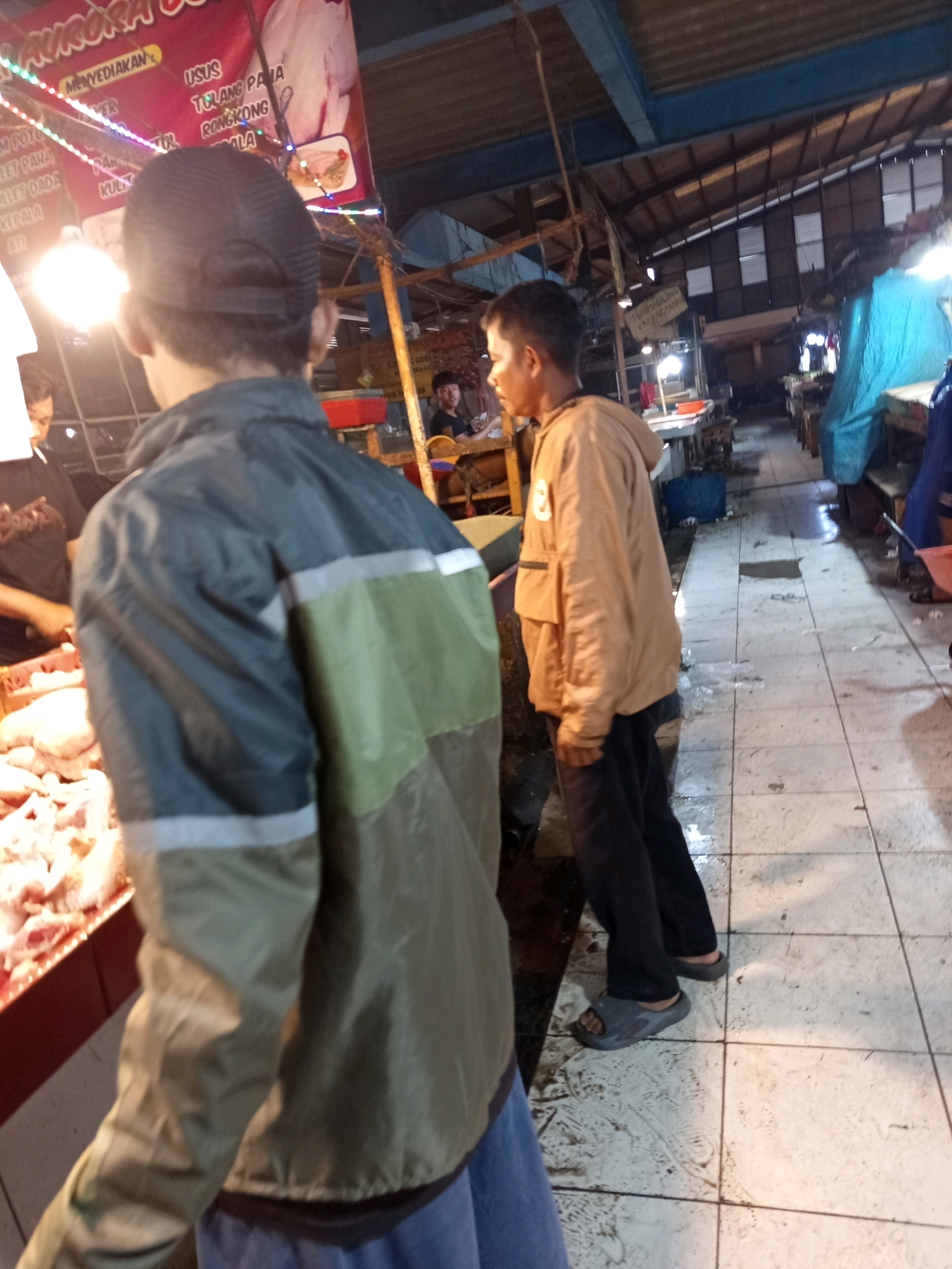 In Karawang, Indonesia, a market scene unfolds with several individuals present. In the foreground, the back of a person wearing a dark green and light olive jacket with reflective stripes is visible. They appear to be standing near a stall displaying raw chicken. To their right, a person wearing a tan jacket and dark pants stands facing away from the camera. In the background, other market stalls are set up under a corrugated roof. A large red banner with Indonesian text is prominently displayed, featuring an image of raw meat. The floor is tiled and appears wet in some areas. The lighting suggests it might be evening or the market is indoors.