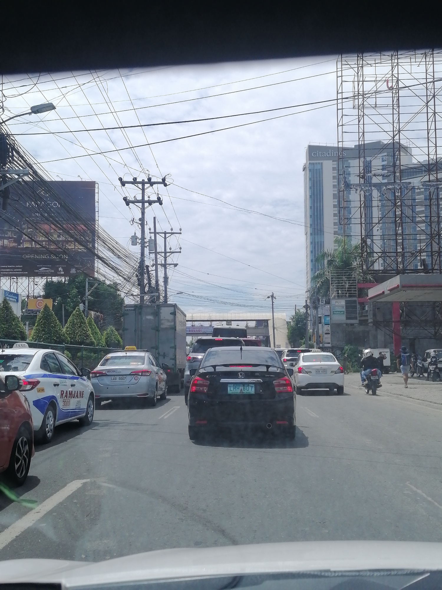 The image depicts a busy street scene in Davao City, Philippines, characterized by a substantial amount of vehicular traffic under a cloudy daytime sky. The perspective is from inside a vehicle, looking ahead at multiple lanes of cars, a truck, and motorcycles.
In the foreground and midground, various vehicles are caught in traffic. A white taxi on the left has "RAMJANE 3088" and "LAR SAB" printed on its side, while another silver taxi ahead of it displays "Krizia" on its roof sign and the license plate "LAO 8007". A black sedan with the license plate "LAH1807" is visible in the right lane, followed by a white car with "KAB 6392". A large light-colored truck is in the central lane. Several motorcycles, including one with a rider and passenger, and a couple of pedestrians are also visible on the right side of the road, near a gas station.
The urban environment is dense with overhead utility wires and cables, which crisscross the sky and are supported by numerous utility poles. On the left, a large billboard advertises "OPENING SOON" and "CEBULANDMASTERS", with fragments like "LEASING", "LIFESE", and "Residencia" also visible. A tall, modern high-rise building with the prominent "citadines" logo on its facade dominates the background skyline. Adjacent to it is a large, empty metal billboard frame. Other visible texts include "TAXI", "AD39", "PUJ", "REGULAR", and a partially visible "Davao" on a distant sign, confirming the location. The scene portrays a bustling urban landscape with typical infrastructure and activity of a major Philippine city.