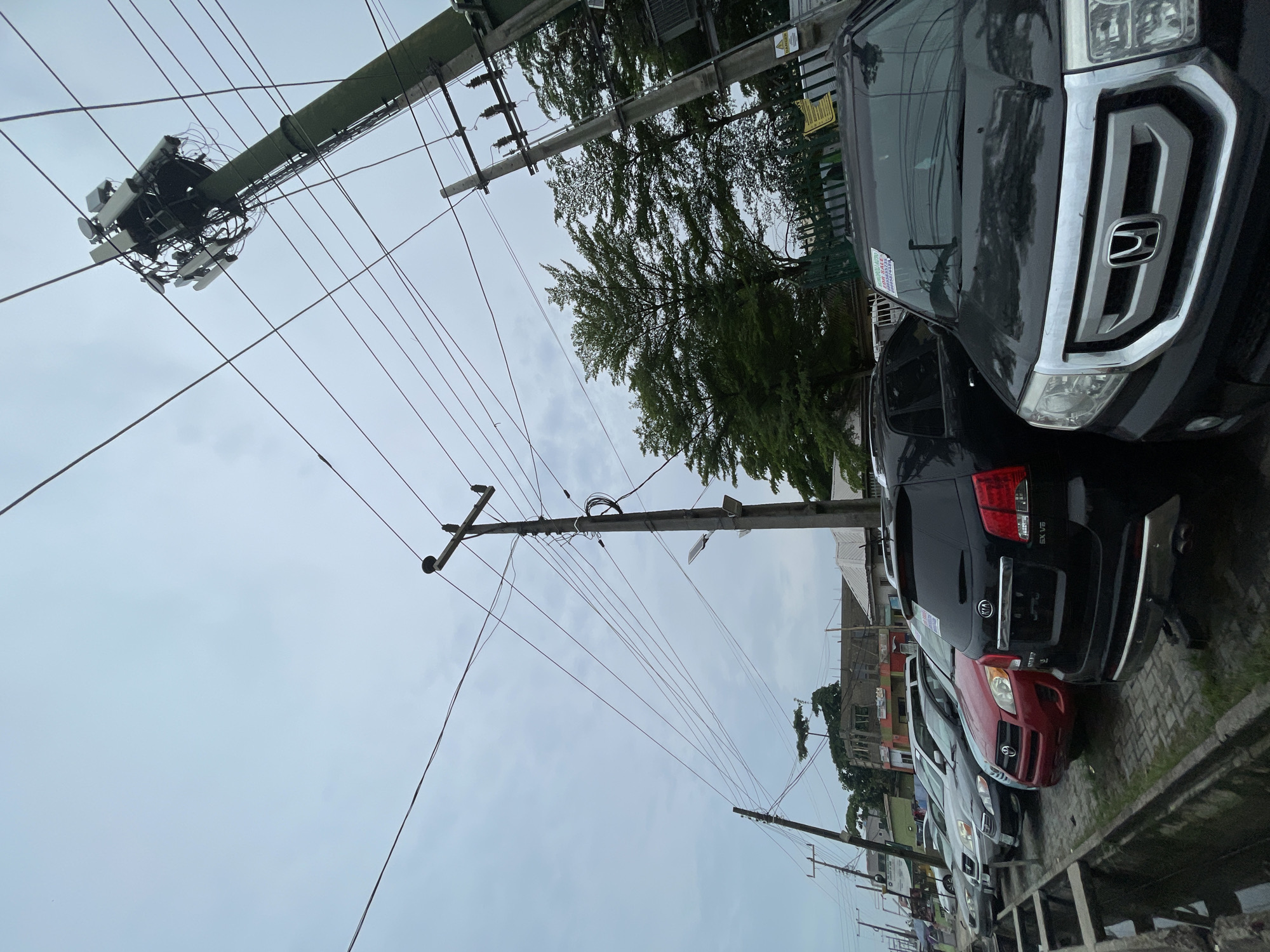 The image shows a street scene in Ado, Nigeria. A row of cars, including black and red SUVs, are parked along the side of a paved road. Overhead, power lines and telephone poles stretch across the frame. A large telecommunications installation is attached to one of the poles. The sky is overcast. Buildings are visible in the background.