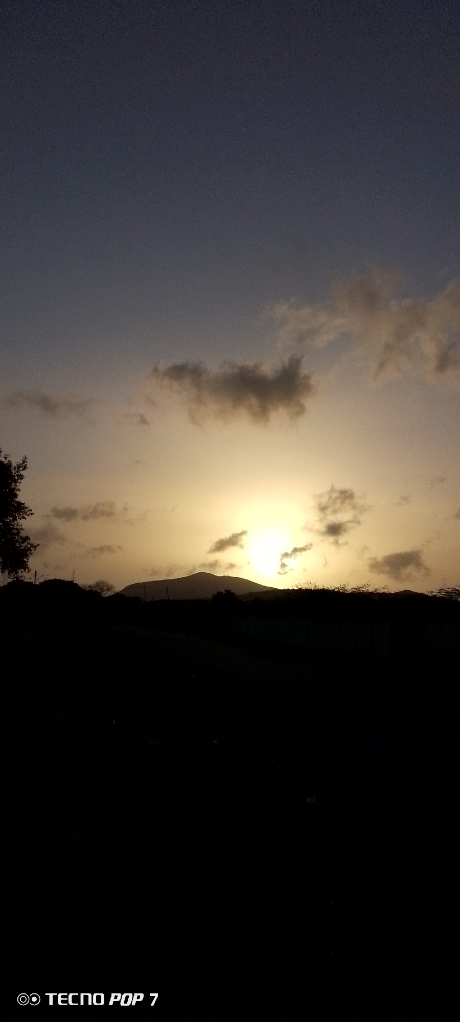 The image depicts a landscape at sunset or sunrise in Altagracia, Venezuela. The sun is partially obscured by clouds and what seems to be a mountain range. The foreground is dominated by a dark, silhouetted area, presumably vegetation or a field. A tree is visible on the left side of the frame. The sky transitions from a darker tone at the top to a brighter, yellow-orange hue near the horizon, where the sun is located. The watermark of a "Tecno Pop 7" phone is at the bottom.