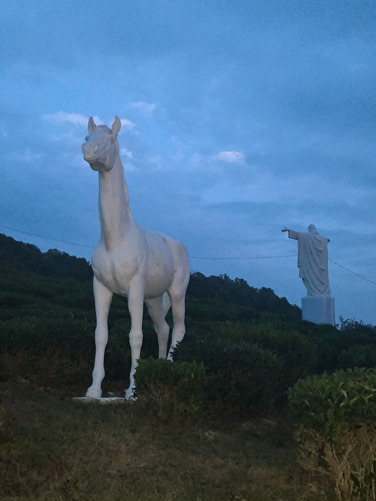 In Kalinovoye Ozero, Russia, at dusk, a large white horse statue stands prominently in the foreground, facing left. The horse is detailed, with a mane, tail, and muscular build, positioned on a grassy slope. Further in the background, to the right and on a higher elevation, a white statue of Jesus Christ with outstretched arms stands on a pedestal. Both statues are set against a twilight sky with scattered clouds and a dark, tree-covered hillside. The scene is illuminated by ambient light, suggesting either early morning or late evening.