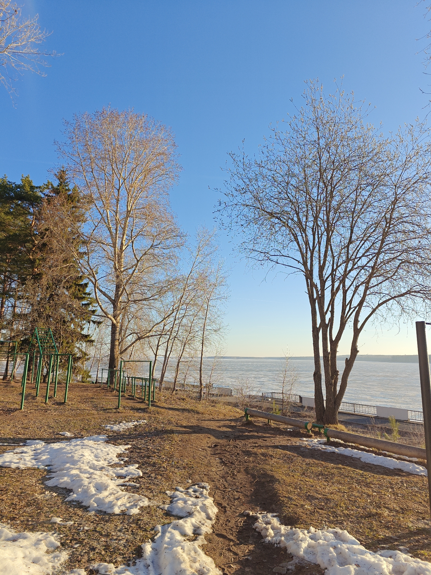 The image depicts a scene in Chaikovsky, Russia, with a clear blue sky above. The foreground features a dirt path with patches of melting snow. To the left, there is a playground area with green metal exercise equipment, partially obscured by trees. Several bare deciduous trees stand prominently, their branches reaching towards the sky. Evergreen trees are visible in the background on the left. In the distance, a body of water is seen, possibly a river or lake, with a white surface suggesting it might be frozen or partially covered with ice. A railing borders the edge of the water, indicating a waterfront area. The lighting suggests it is daytime, likely in early spring or late winter, given the mix of bare trees and snow.