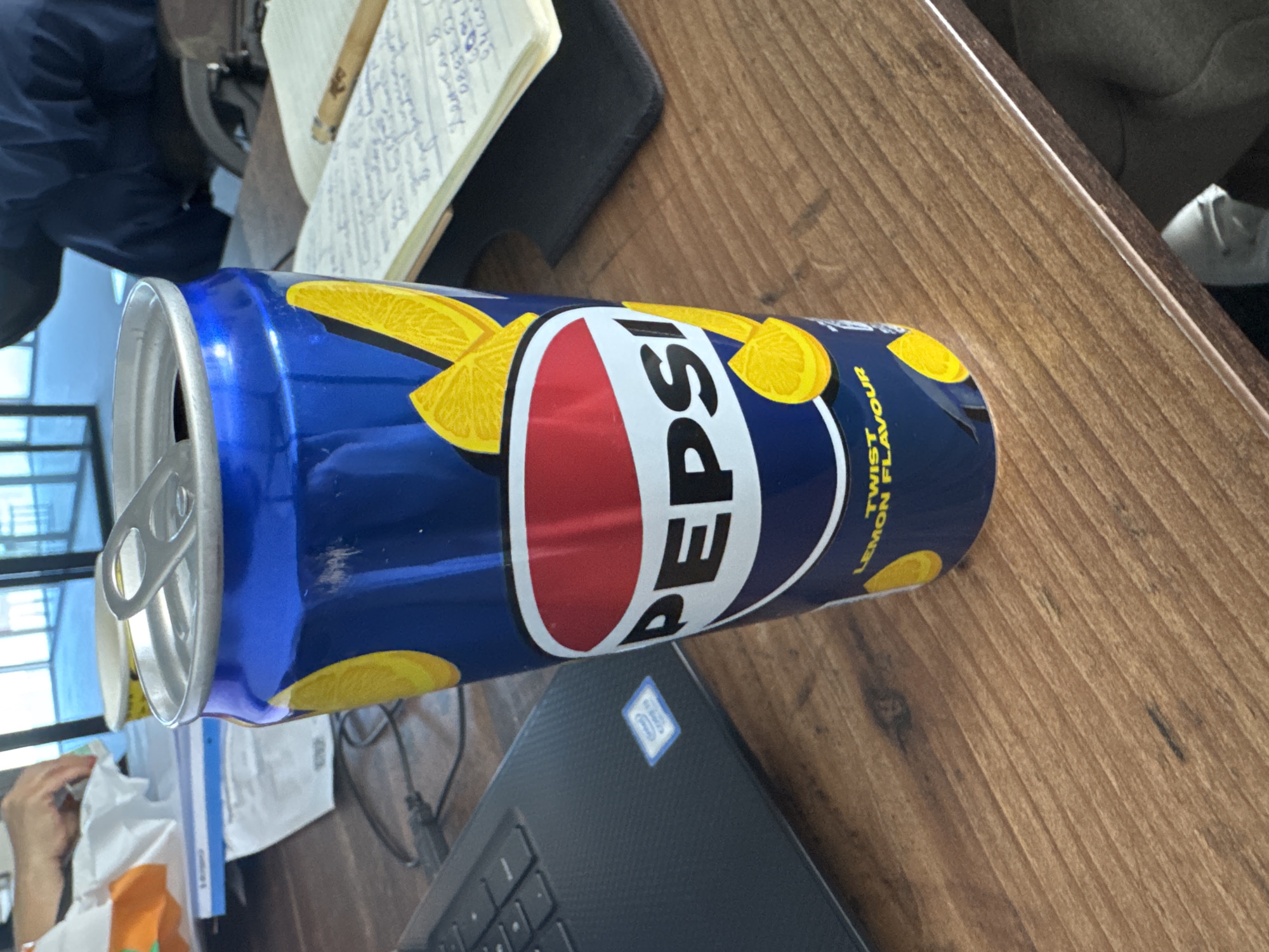 A slightly canted, eye-level indoor shot features a can of Pepsi Twist Lemon flavor in the foreground. The can is primarily blue, with yellow lemon slice graphics and the white "PEPSI" logo in a red and white oval. The can's silver top is visible, with the pull tab extended.
The can sits on a wooden surface, possibly a desk or table, with a light-colored wood grain pattern. A black laptop with a visible Intel logo is partially visible beneath the can. To the left of the can is a notebook, a pen, and a glimpse of a person's hands and arm wearing an orange garment.
In the background, through a window, a grid of blue sky, possibly reflecting the cityscape or a nearby building. It's difficult to confirm that this photo was taken in Sofia, Bulgaria, because there are no identifying visual cues.