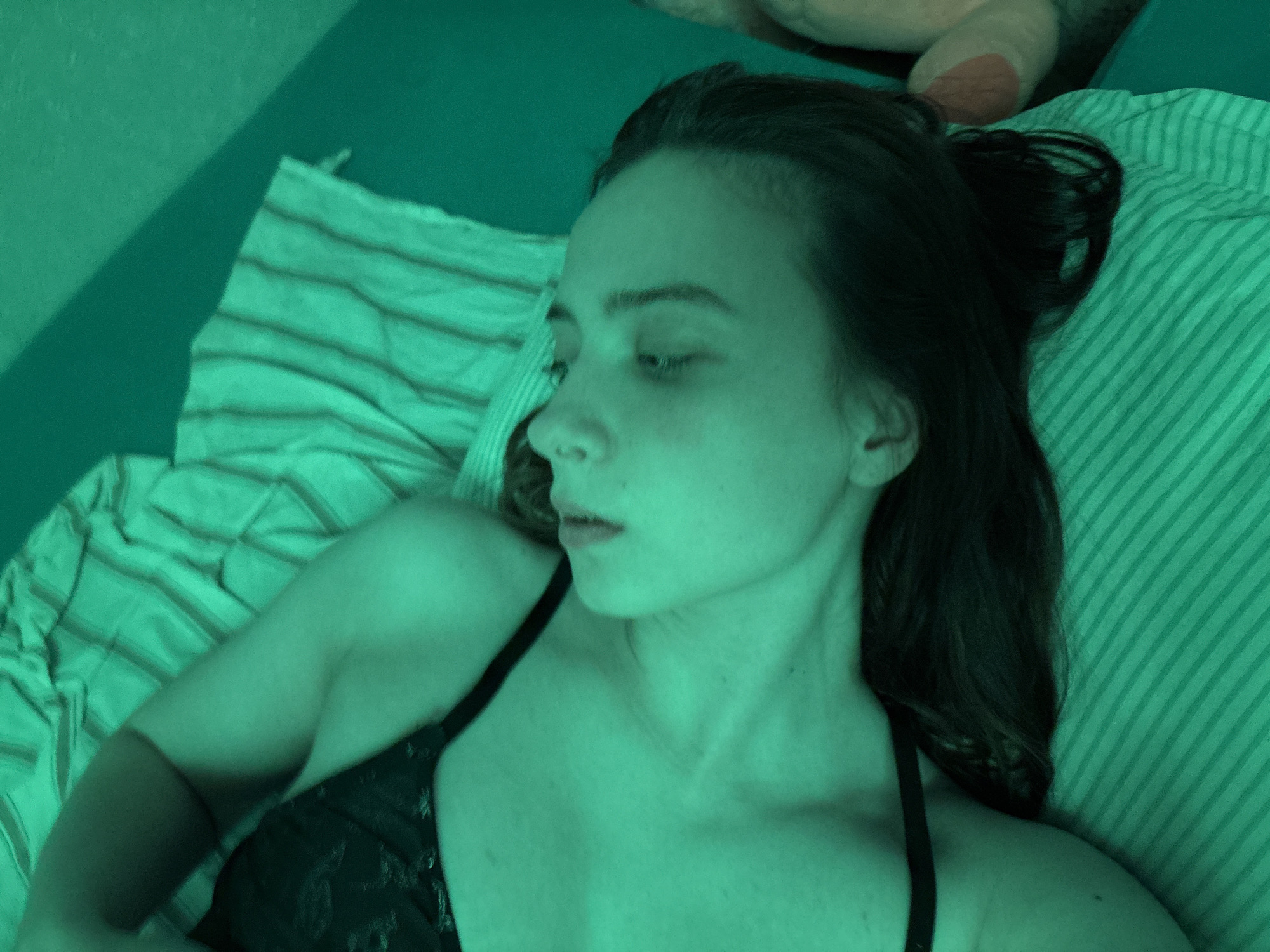 A young woman is depicted lying on a bed in an indoor setting, illuminated by a strong green light that bathes the entire scene in a vibrant green hue. She is shown in profile, facing slightly to the left with her eyes partially closed or looking down, conveying a pensive or relaxed mood. Her dark hair is loose, resting against a striped pillow that matches the green tone of the room.
She is wearing a dark-colored top, possibly a bra or camisole, with thin straps visible on her shoulders. The bedding features a pattern of lighter green stripes on a darker green background. A wall, also in a green shade, forms the background, with a distinct shadow cast on its upper left portion. In the upper right corner, a portion of a light-colored plush toy is partially visible. The overall ambiance is intimate and moody due to the specific lighting. No visible text is present in the image. The location context is Прикамский, Russia.