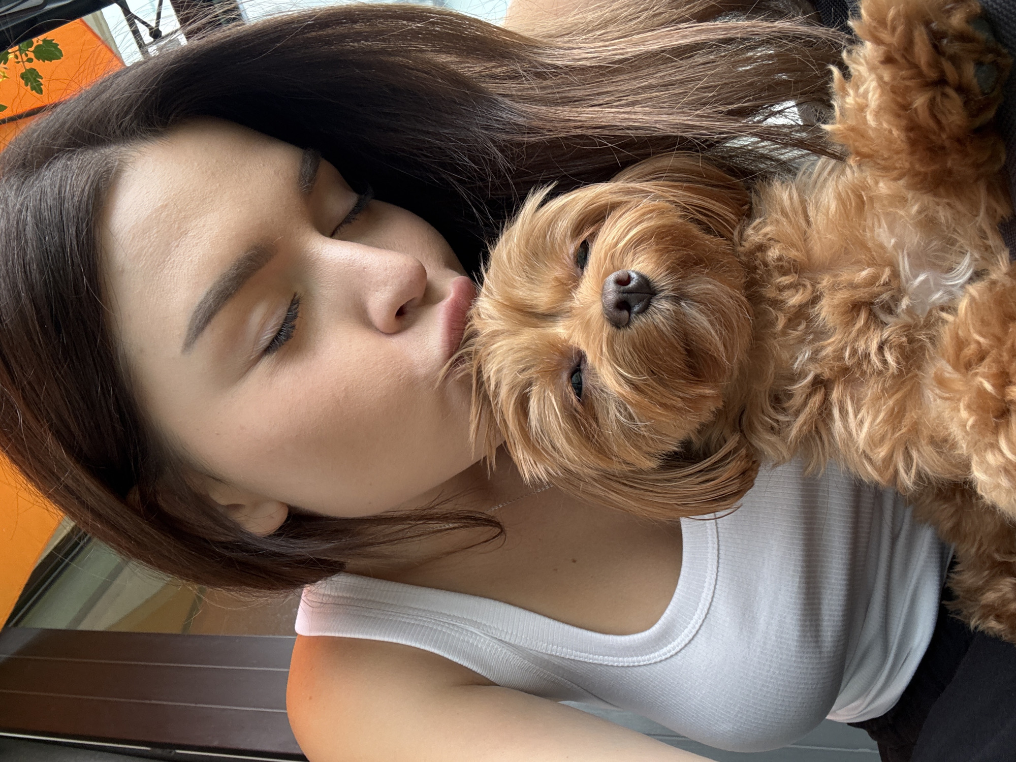 The image shows a close-up of a young woman with her eyes closed, kissing a small, light brown dog. The woman has long, dark brown hair that covers part of her face and the dog. She is wearing a white tank top. The dog has fluffy, curly fur and is resting its head on the woman's shoulder. The background is out of focus but appears to be an indoor setting with natural light. The overall mood is affectionate and tender. There is no visible text in the image.