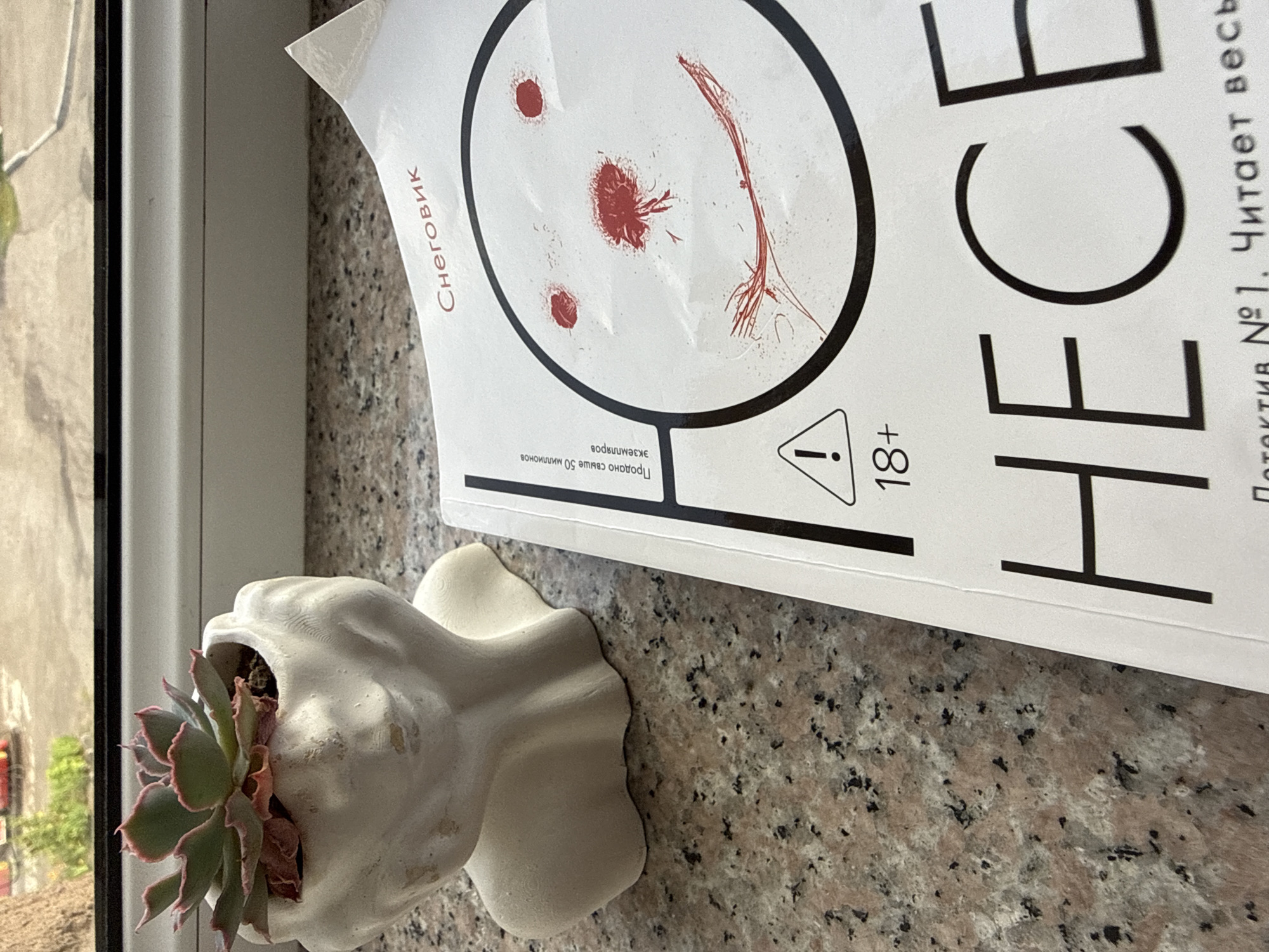 The image depicts a close-up, high-angle shot of a book cover placed on a speckled granite countertop. To the left of the book is a white, abstract bust-shaped planter containing a succulent plant. The book cover is white with black and red text and graphics. Prominently featured is a drawing of a snowman's face with red markings for eyes, nose, and mouth, suggesting blood. The text on the cover includes "Снеговик" (Snowman) in red at the top right, "Продано свыше 50 миллионов экземпляров" (Over 50 million copies sold) in small black text on the left, and "18+" next to a warning triangle graphic. Large black letters spell out "НЕ CE" at the bottom, suggesting the title of the book is incomplete in this view. The environment appears to be indoors, possibly near a window, with the granite countertop being the primary surface. The city of Sevan, Armenia, is not depicted or suggested by any elements in the image.