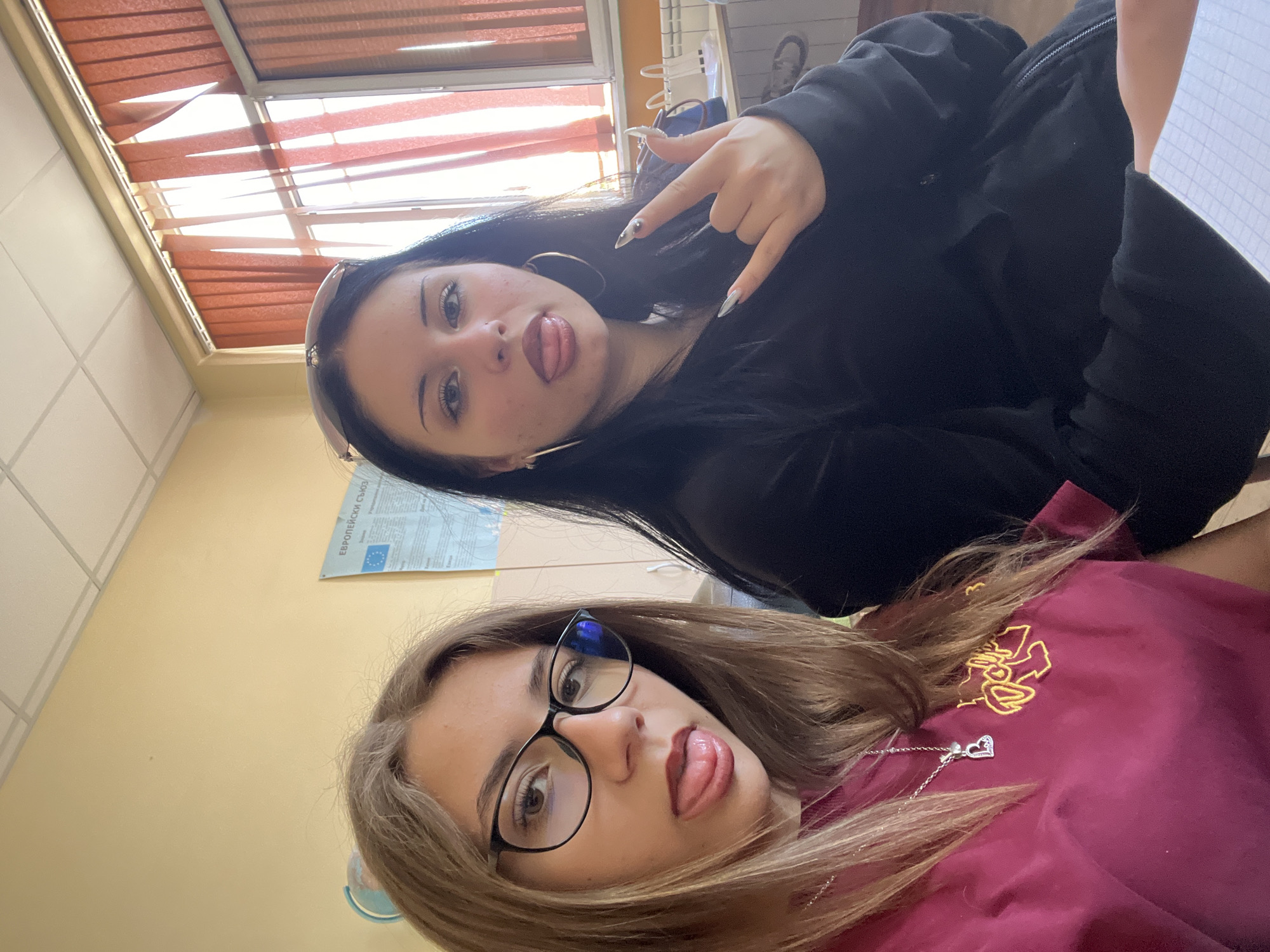 Two young women pose for a selfie indoors, possibly a classroom. The woman in the foreground has light brown hair, wears glasses, and a maroon shirt with an unknown logo. She is sticking her tongue out. The woman in the background has long, dark hair, large hoop earrings, and wears a black hooded sweatshirt. She is sticking her tongue out and making a rock and roll hand gesture.
The background includes a window with blinds, light yellow walls, and a poster in Bulgarian, partially reading “Европейски съюз.” This suggests the location is in Bulgaria, and possibly a school within the city of Plovdiv given the prompt.