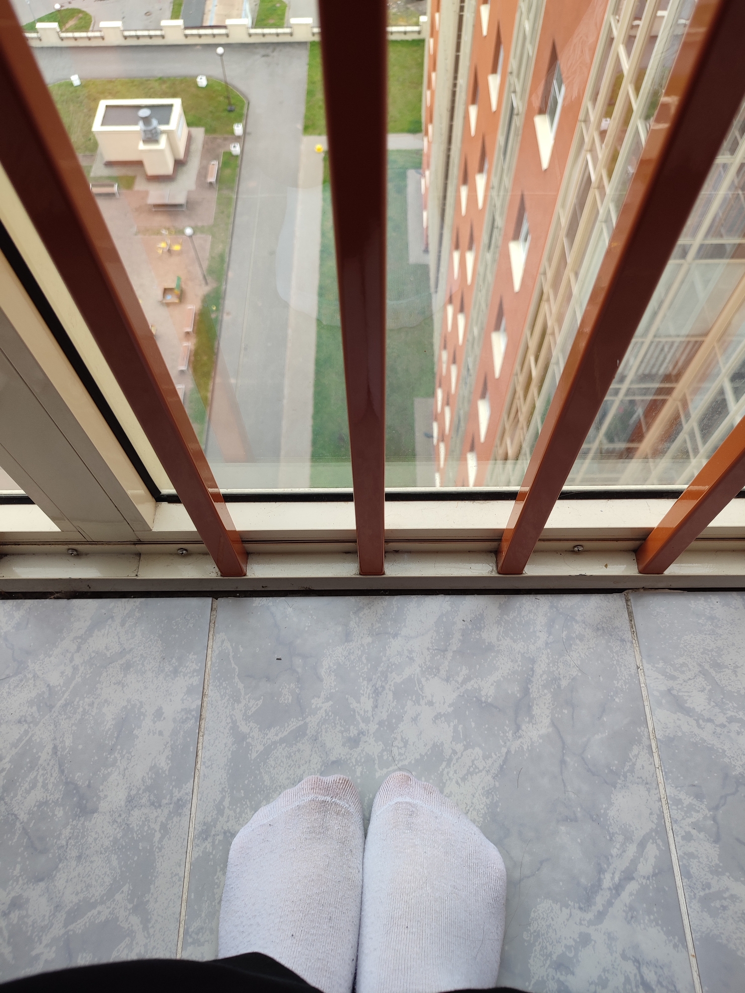 The image is a first-person perspective of someone standing on a balcony in Murino, Russia. The foreground shows the person's feet wearing white socks, standing on a grey tiled floor. The balcony railing, with brown vertical bars and a white horizontal frame, is visible. Beyond the railing, a large, orange apartment building with many windows dominates the view. Below the balcony, a courtyard area is visible, featuring a small building, benches, a playground with yellow structures, and a paved pathway. The overall environment suggests an urban residential setting.
