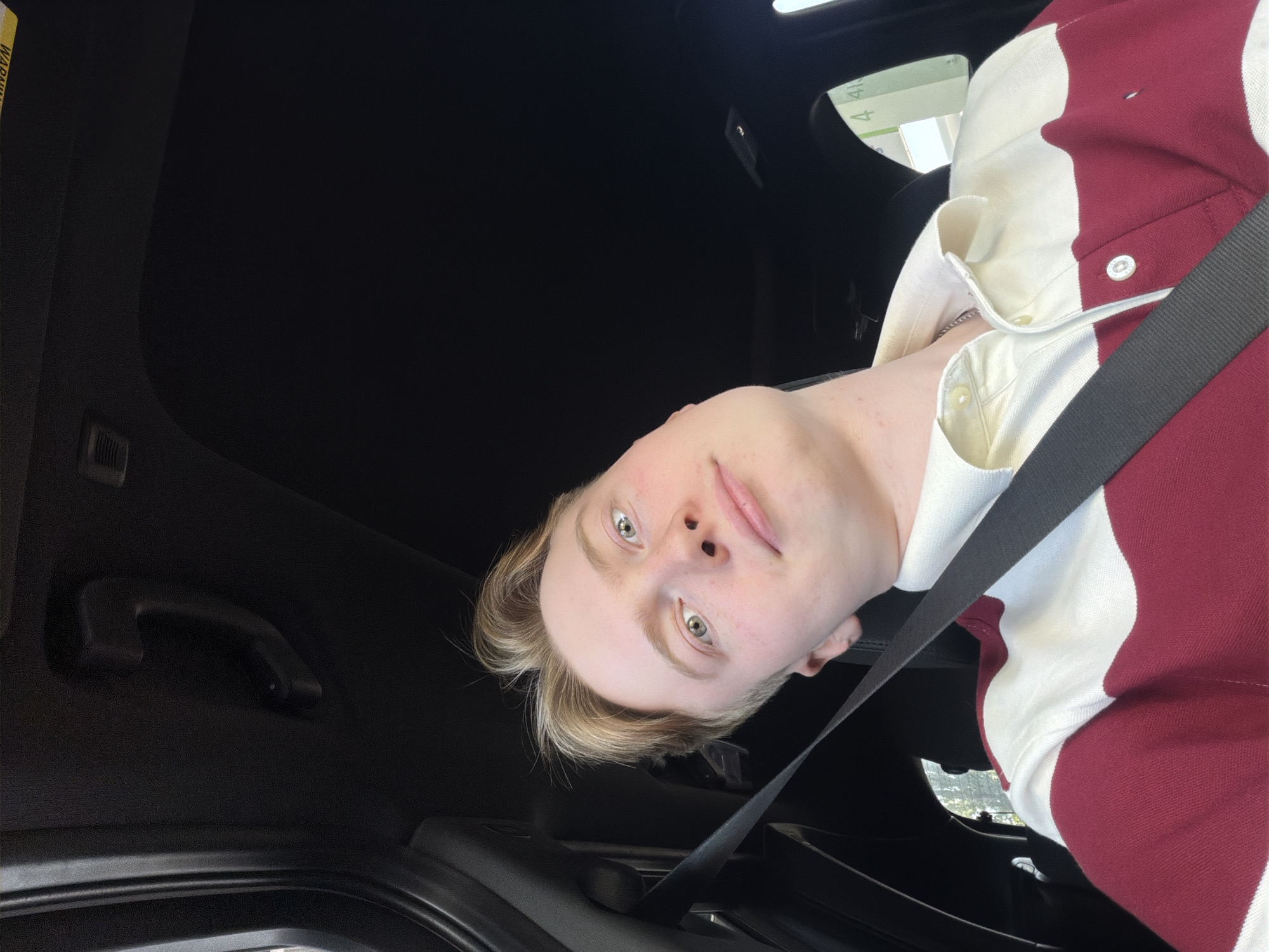 A young Caucasian male with blond hair and light green eyes is taking a selfie from the driver's seat of a car. He is wearing a red and white striped polo shirt and a black seatbelt. The interior of the car is dark, with a black headliner and a black door panel visible. Outside the car, a bright, sunny sky is visible, as well as what appears to be a gas station with green signage. The setting is indeterminate but could be a city in Ukraine.