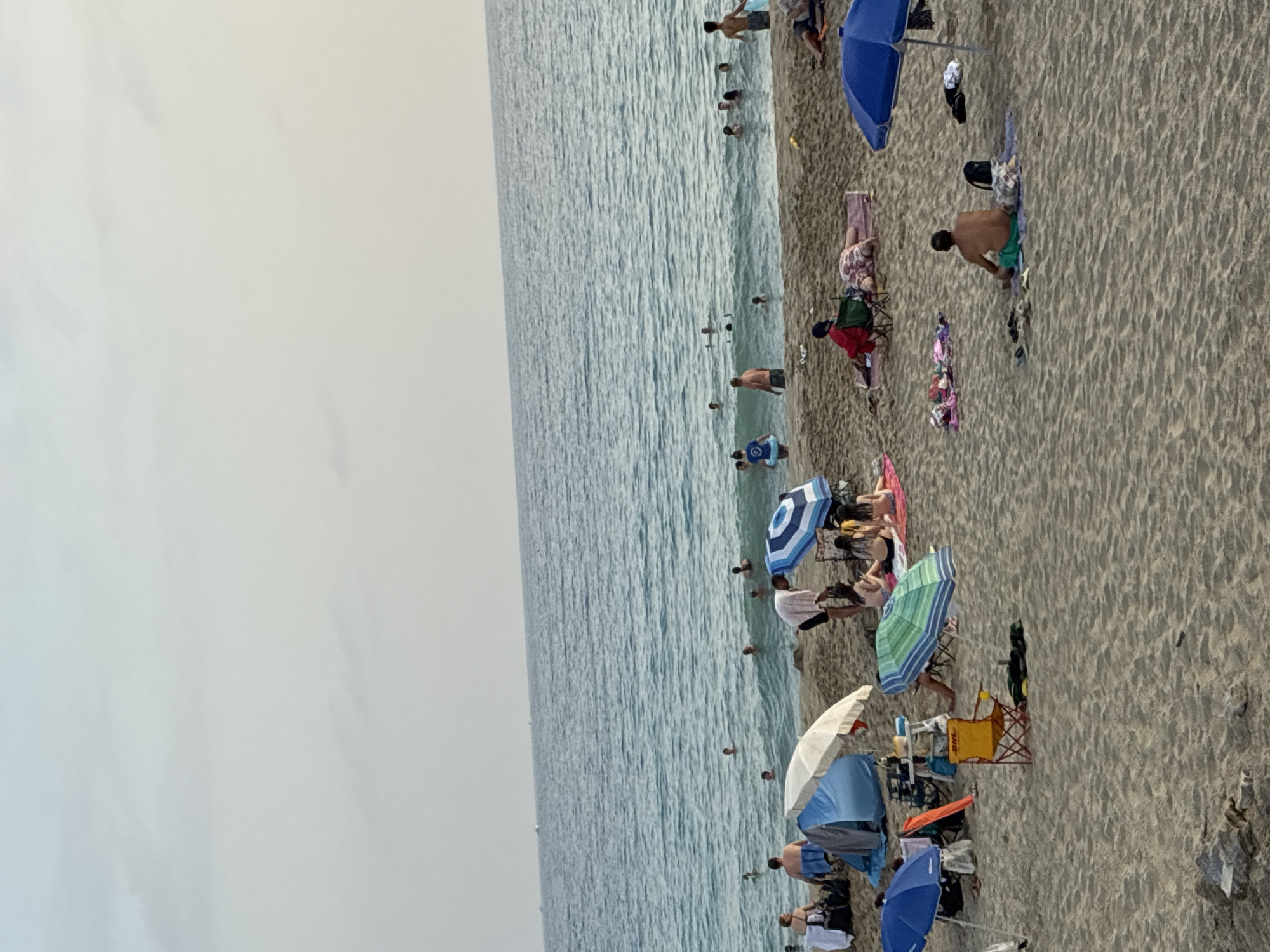 The image depicts a crowded beach in Alanya, Turkey. The sea is a vibrant blue with gentle waves, and numerous people are swimming and wading in the water. On the sandy shore, beachgoers are relaxing under umbrellas, sitting on chairs, or lying on towels. Various beach equipment, including tents, chairs, and bags, are scattered across the sand. The overall scene is one of a typical summer day at a popular tourist destination.