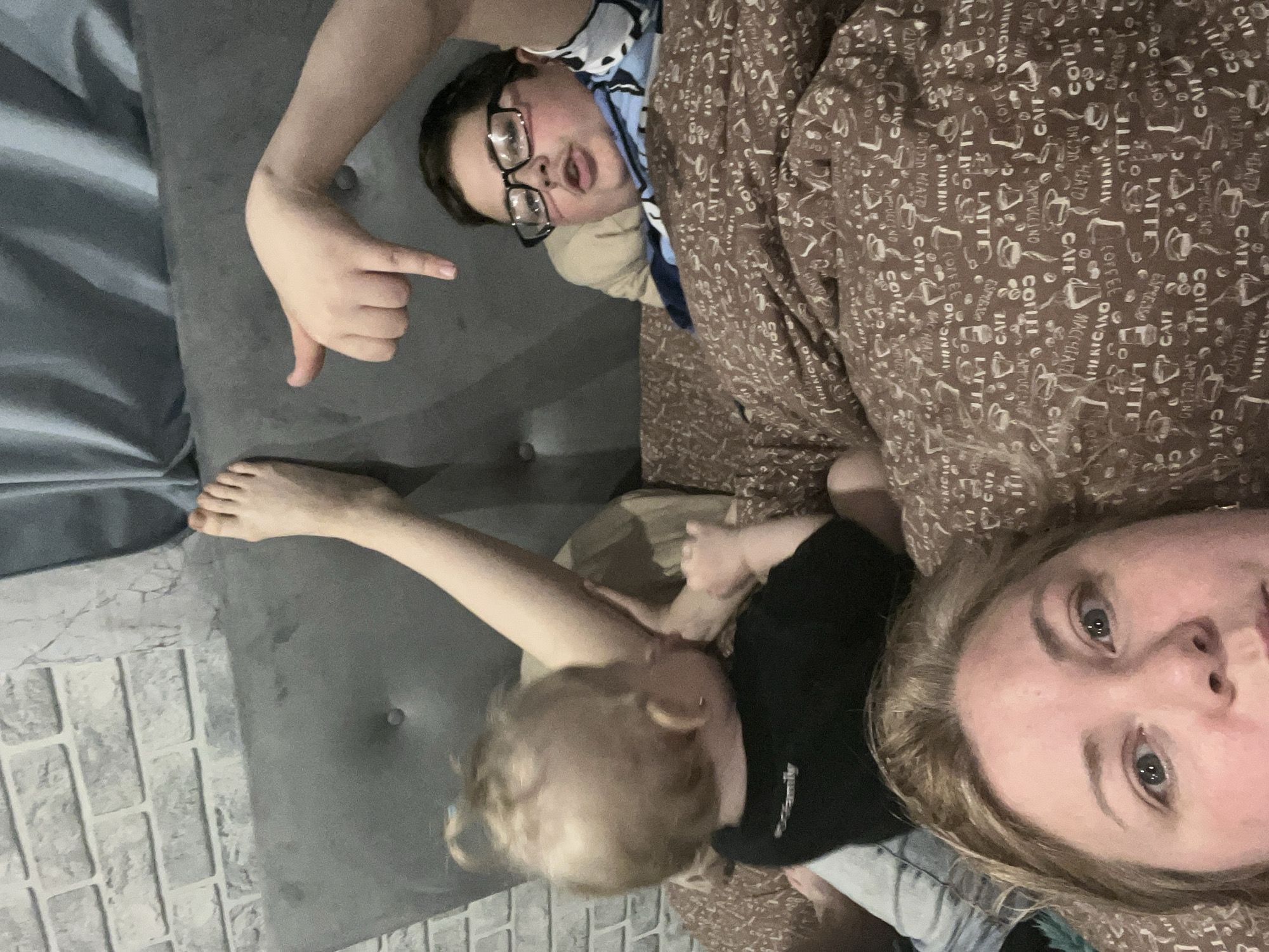 Three individuals are visible in the image, likely a family group, possibly in a domestic setting. A child with blonde hair appears to be lying on or near a gray sofa. An adult, possibly the mother, with blonde hair and fair skin, is also visible in the foreground, close to the camera, and appears to be part of the scene. An older child or young adult wearing glasses is partially covered by a coffee-themed printed blanket, pointing toward the gray sofa. The background includes a gray wall with a stone pattern. The image appears to be a casual snapshot taken indoors, with the subjects lying down or relaxing on the sofa. There is no specific evidence to indicate the location is Vesyoloye, Russia, and this information cannot be confirmed.