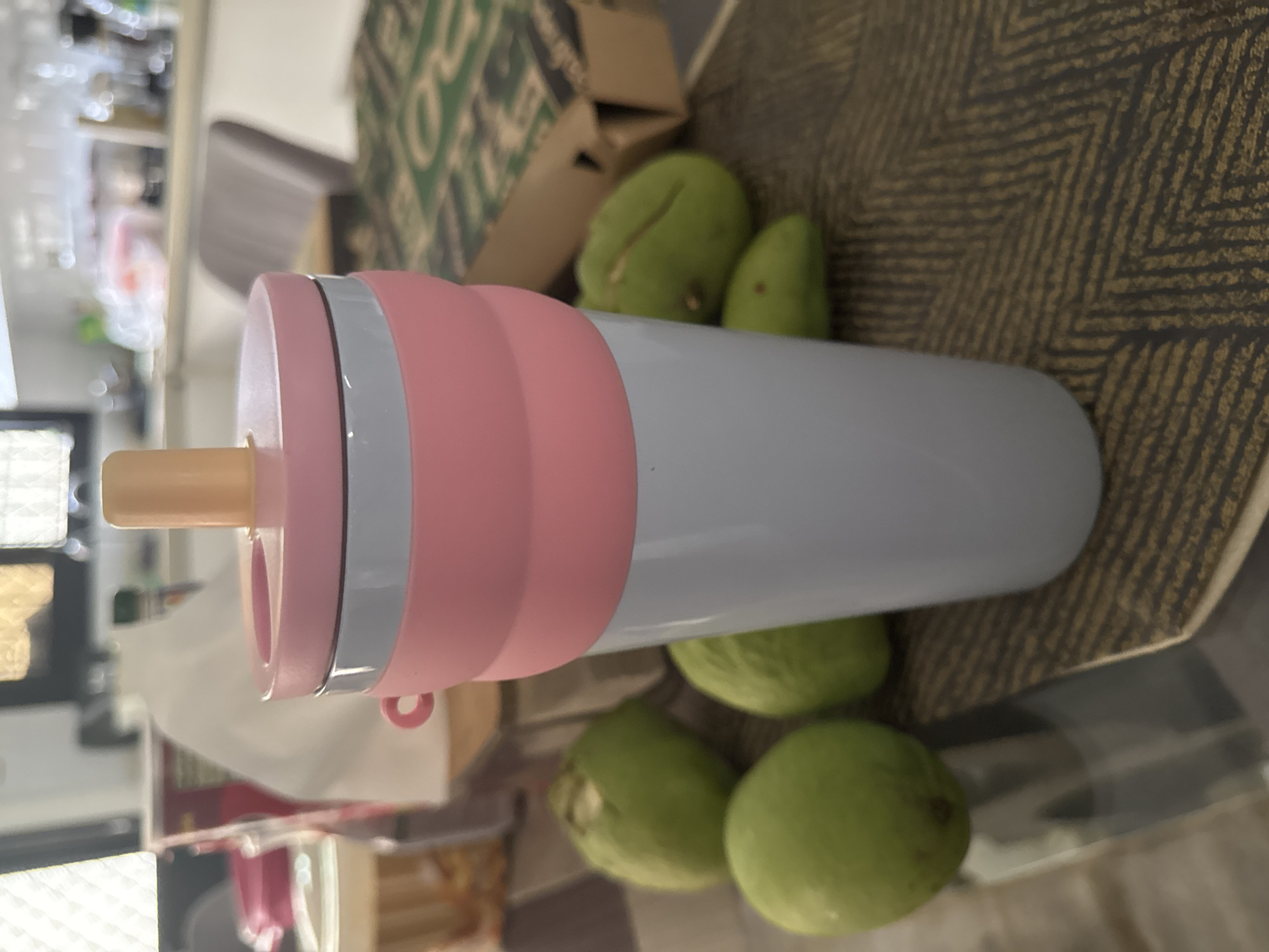 The image displays an indoor scene in Davao City, Philippines, centered on a reusable, collapsible drinking cup and several green fruits. The cup, positioned prominently in the foreground, consists of a light grey base, a pink accordion-style silicone mid-section featuring a small pink loop handle on its left, a thin grey band, and a flat pink lid from which a cylindrical beige straw or spout emerges. Five ovoid green fruits, consistent with unripe mangoes, are scattered on a dark brown surface with a subtle, lighter brown zigzag pattern, surrounding the cup's base. In the blurred background to the left, various containers and white packaging are visible, alongside a window with a white grid pattern. To the right of the cup, a brown cardboard box, featuring a partial green logo, rests on the patterned surface. The distant background reveals a blurry domestic setting, likely a kitchen or dining area, with light-colored countertops, fixtures, and a grey upholstered chair, indicating a residential environment. No individuals or specific actions are depicted.
