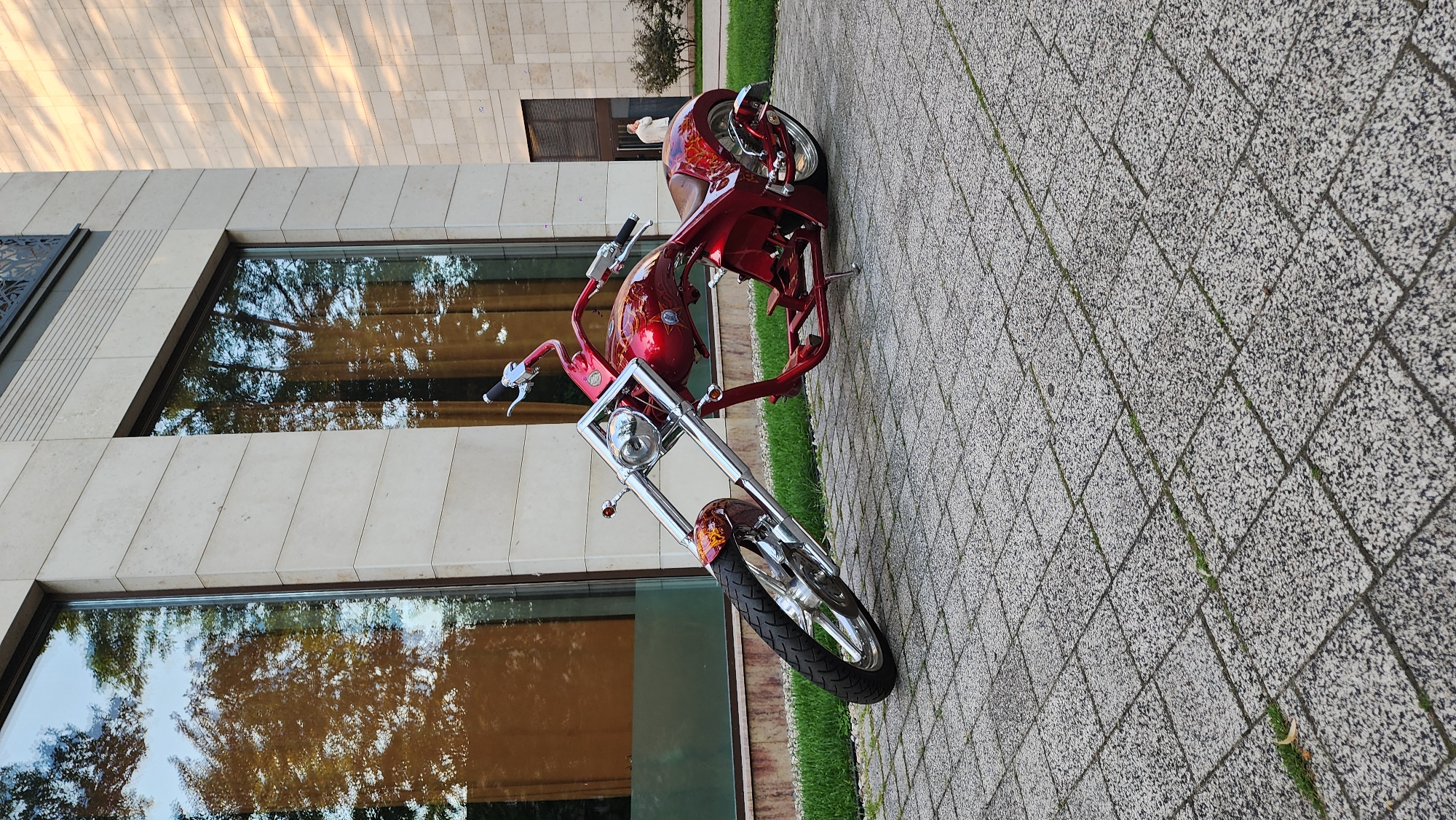 A custom chopper motorcycle, painted in a vibrant red with flame detailing, is parked on a paved walkway outside a modern building. The motorcycle is the primary subject, with its chrome accents and intricate paintwork prominently displayed. The scene is set during the day, with the sun casting long shadows across the tiled ground, suggesting late afternoon or early morning light. The building behind the motorcycle features large, reflective windows that mirror the surrounding trees and sky, creating a sense of depth and blending the man-made structure with the natural environment. The walkway is paved with square, speckled tiles, and patches of green grass are visible between them. There is no text visible in the image. The overall impression is one of a still, observational shot of a striking vehicle in an urban or suburban setting.