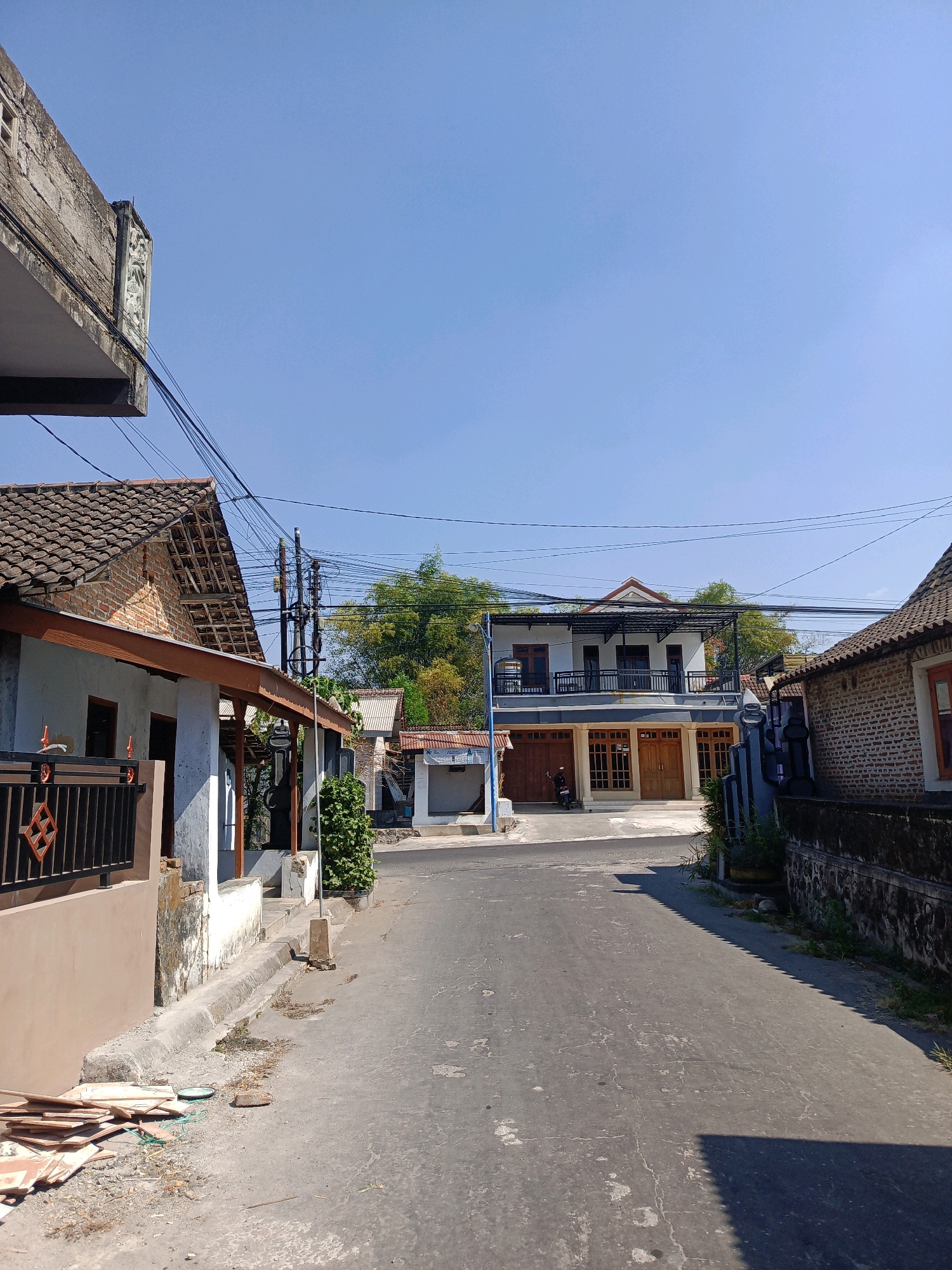 The image depicts a street scene in Tamanan, Indonesia, under a clear blue sky. The road surface is asphalt, showing signs of wear and tear with some discoloration and cracks. On the left side of the road, a light-colored building with a tiled roof and a dark-colored fence is visible. Adjacent to it, another building with a brick facade and a tiled roof is partially seen. Further down the street, a two-story building with multiple doors on the ground floor and balconies on the upper floor stands prominently. A motorcycle is parked in front of this building. On the right side of the road, a brick wall and a building with a tiled roof are present. Overhead, numerous electrical wires are strung across the scene, supported by poles. Green foliage, including trees and plants, is interspersed throughout the background, suggesting a semi-urban or suburban environment. Debris, including wooden planks and a green rope, is scattered on the side of the road near the building on the left. The overall impression is a quiet, daytime street in a residential area.